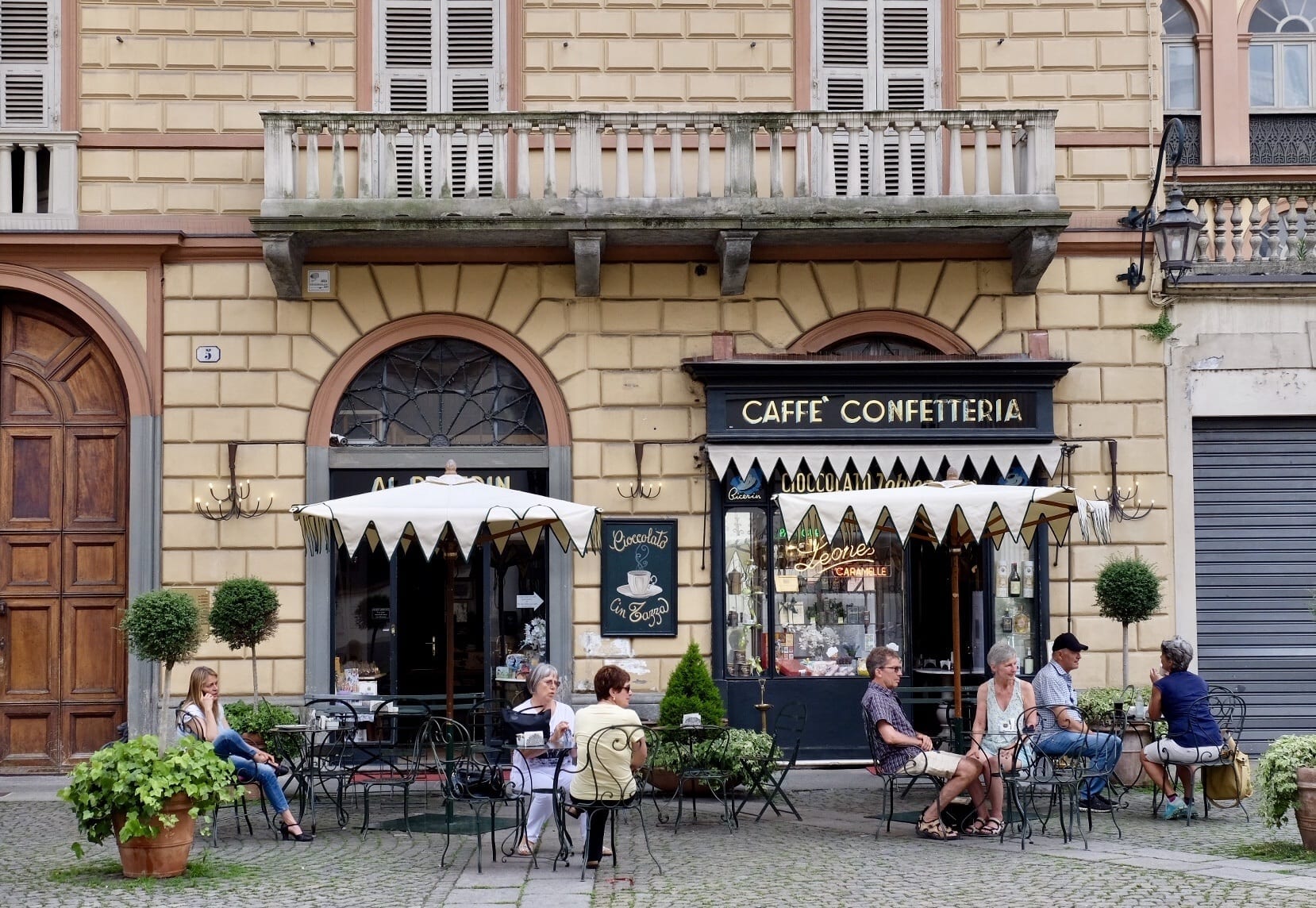 Several people sit at a sidewalk cafe in Torino, Italy. There are umbrellas and the building has a balcony on it.