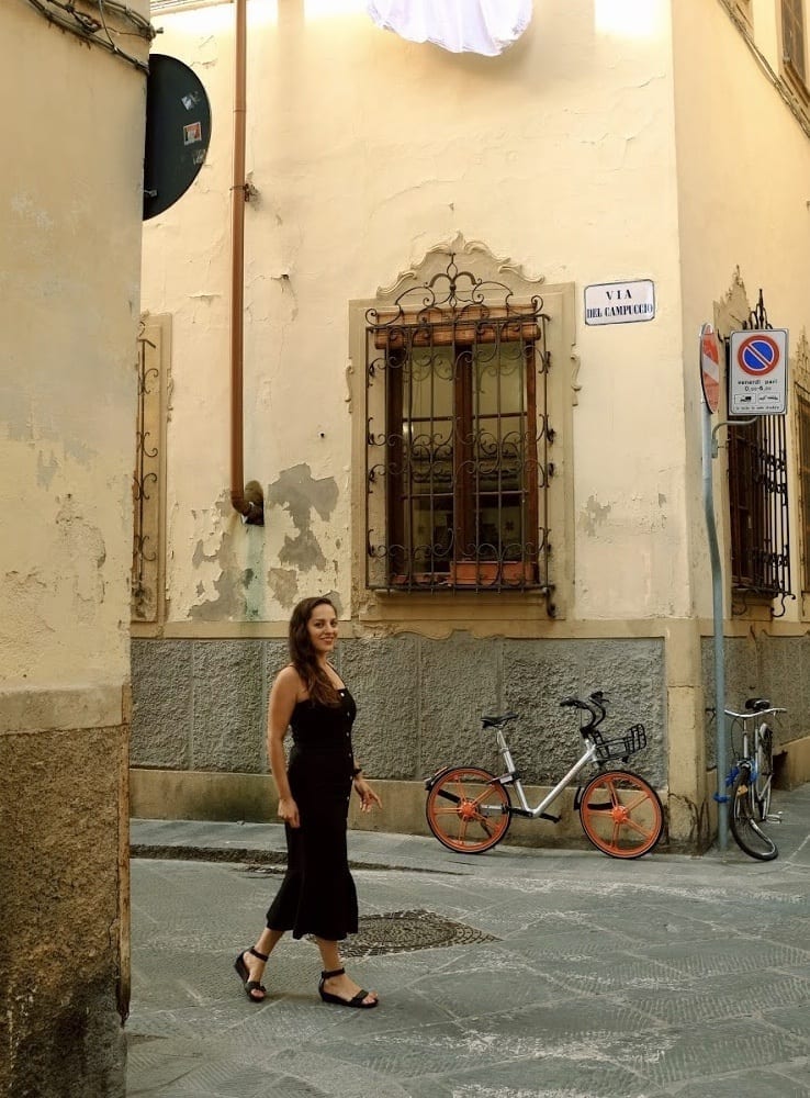 Kate walks down a side street in Florence wearing a long black midi dress with a flared bottom. There is a bike and a wooden window with iron bars on it behind her.