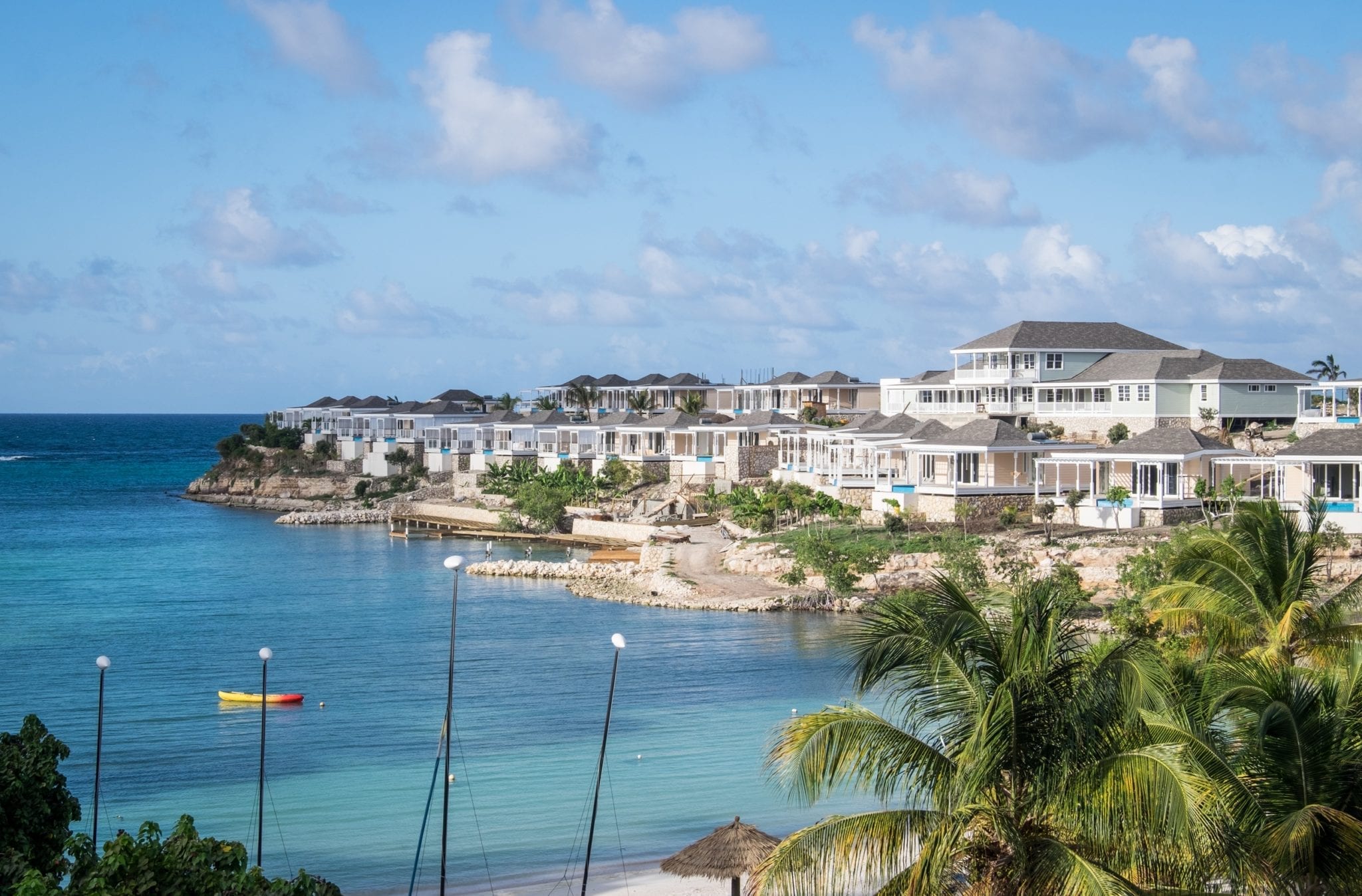 A view over the turquoise Caribbean Sea with white hotel rooms on a piece of rocky land to the right. There are palm trees in one corner.