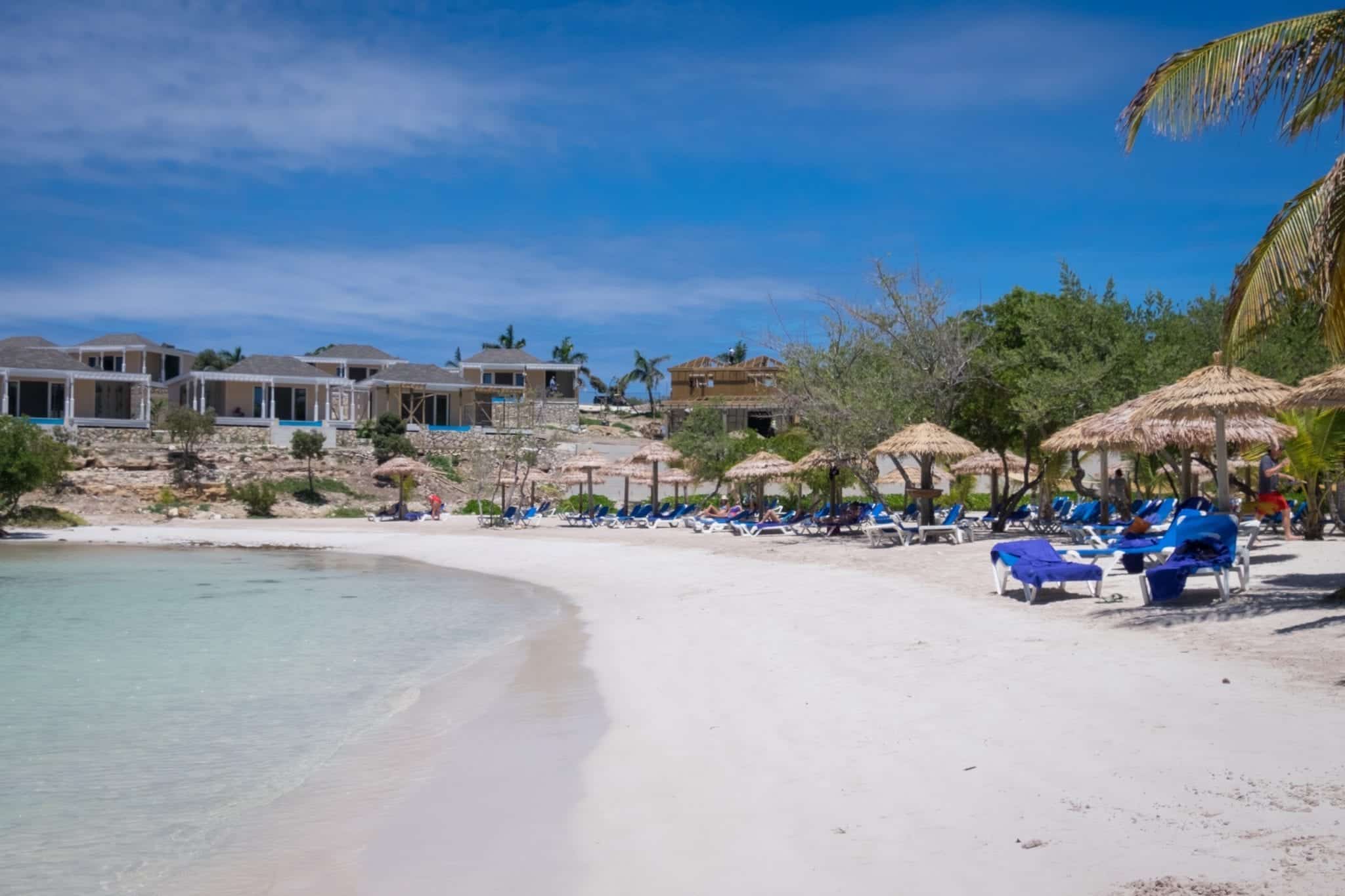 A long turquoise beach with beach chairs underneath umbrellas lined along the beach. The water is nearly clear.