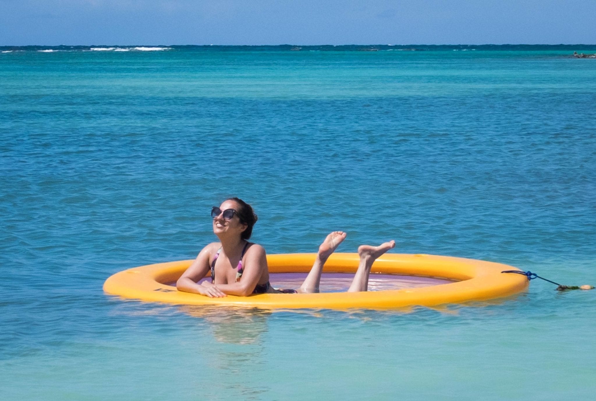Kate is in a yellow "water hammock" -- a round hammock-like device in the middle of the turquoise Caribbean Sea underneath a blue sky with a few clouds. Kate is wearing sunglasses and is on her elbows with her legs playfully in the air.
