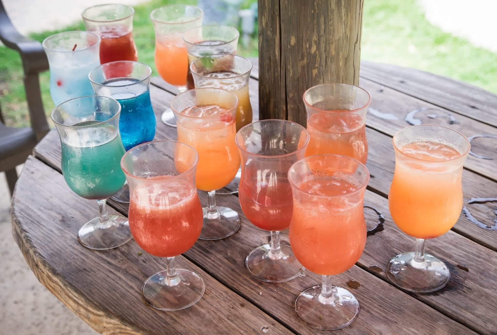 A wooden table topped with 13 various brightly colored cocktails in water goblets, some of them more finished than others.