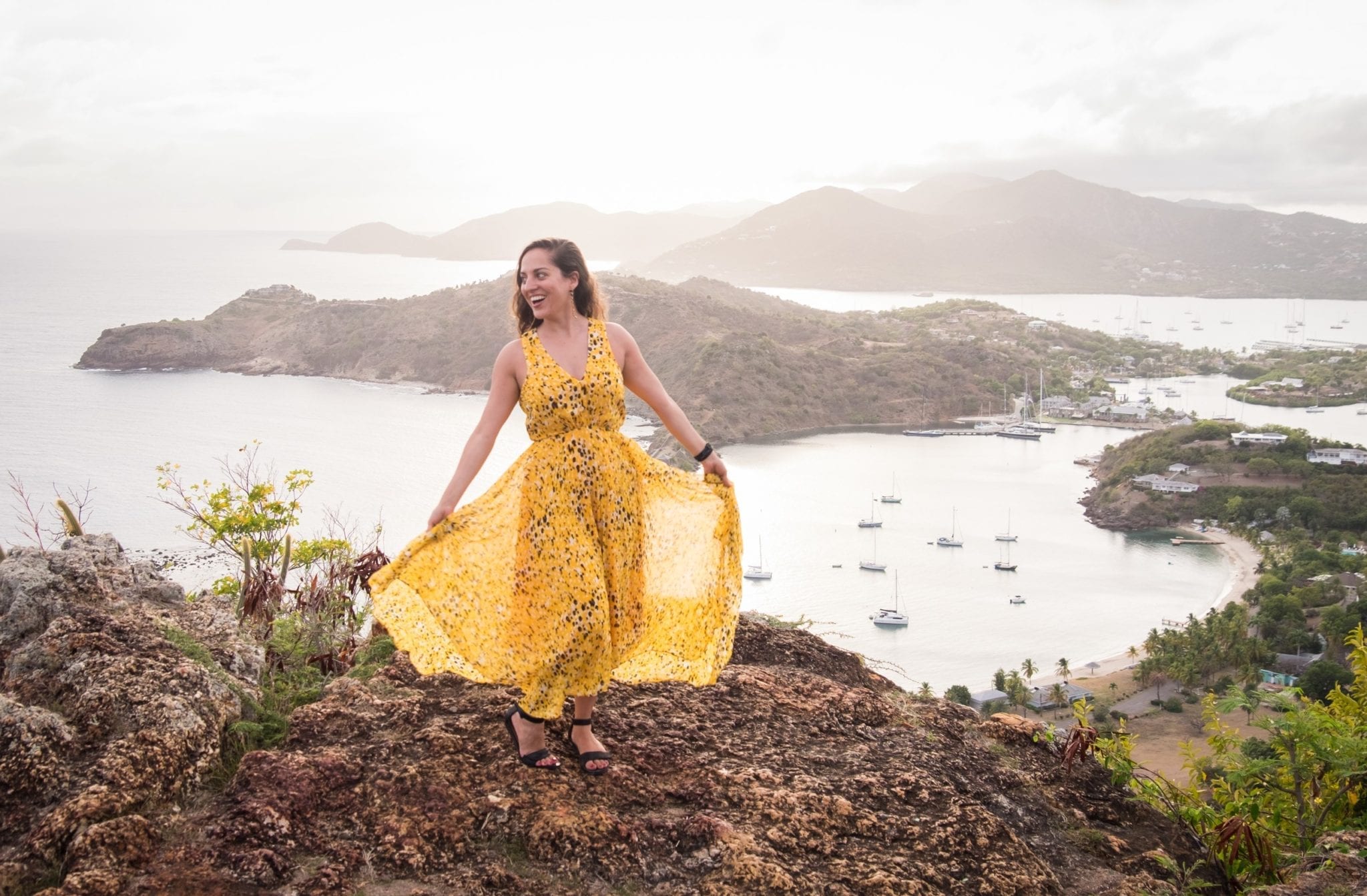 Kate wearing a long yellow and black-spotted gauzy gown over an overlook in Shirley Heights, Antigua. Kate is holding the gauzy outer layer open and smiling. In the background is a view over the White Sea down below, and mountains in the distance.