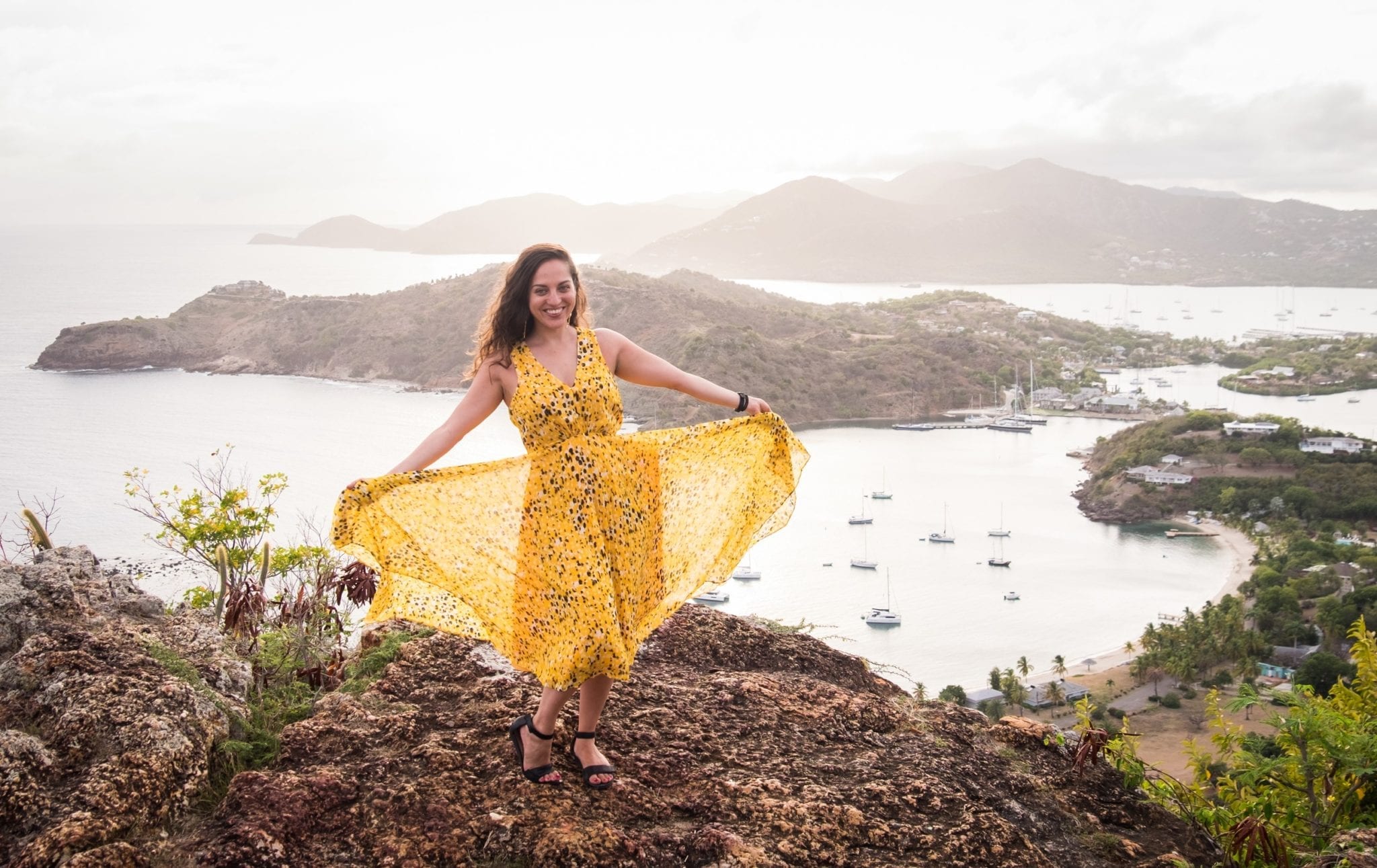 Kate wearing a long yellow and black-spotted gauzy gown over an overlook in Shirley Heights, Antigua. Kate is holding the gauzy outer layer open and smiling. In the background is a view over the White Sea down below, and mountains in the distance.