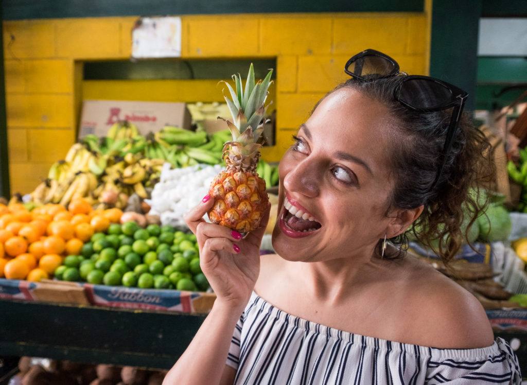 Kate holds a tiny Antigua black pineapple in her hand. It is the size of her fist. She holds it next to her mouth and pretends to eat the whole thing, a smile on her face. In the background are displays of fruit, including limes and oranges.