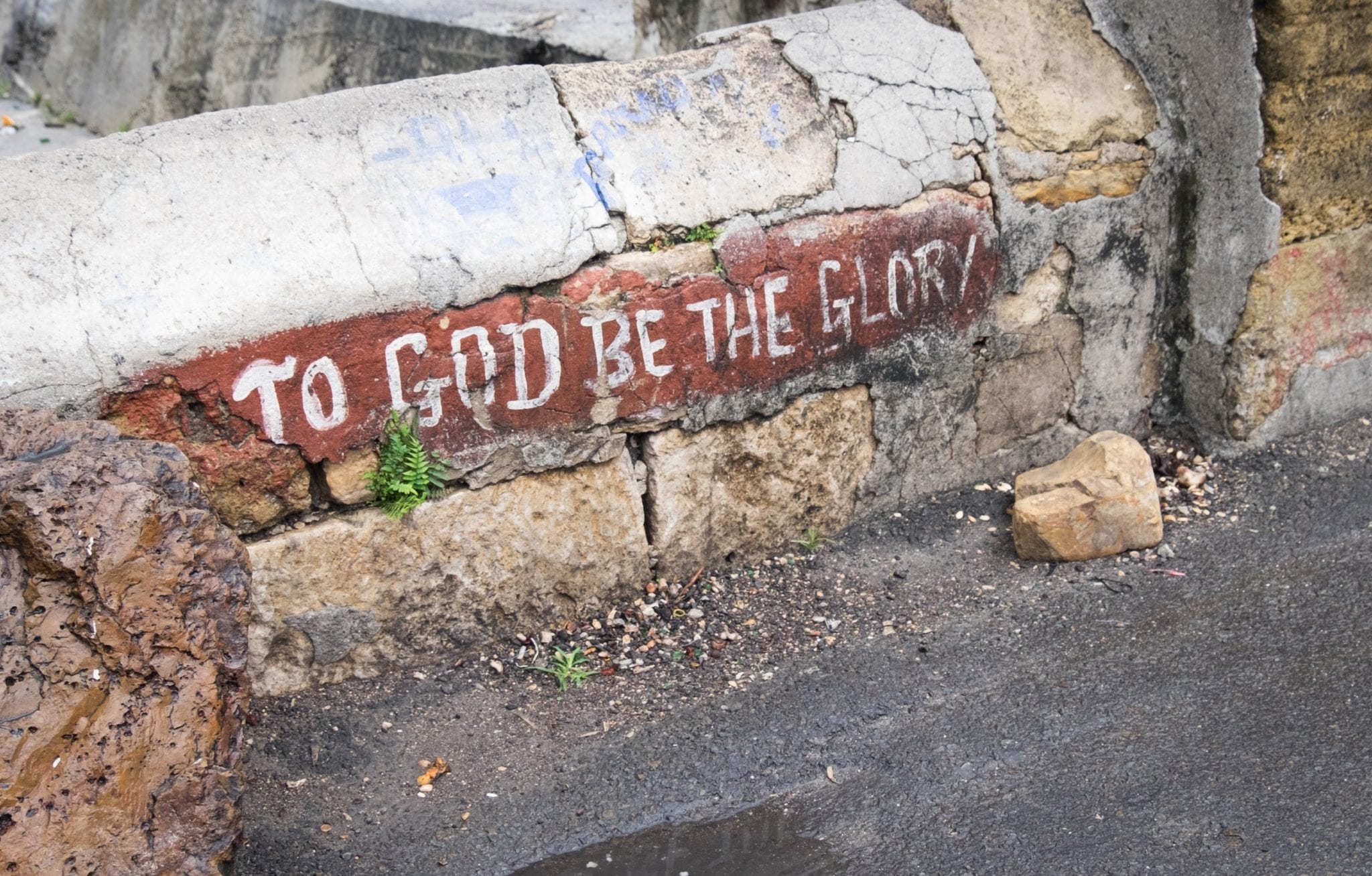 A cracked wall on a sidewalk reads "To God Be the Glory" in St. John's Antigua