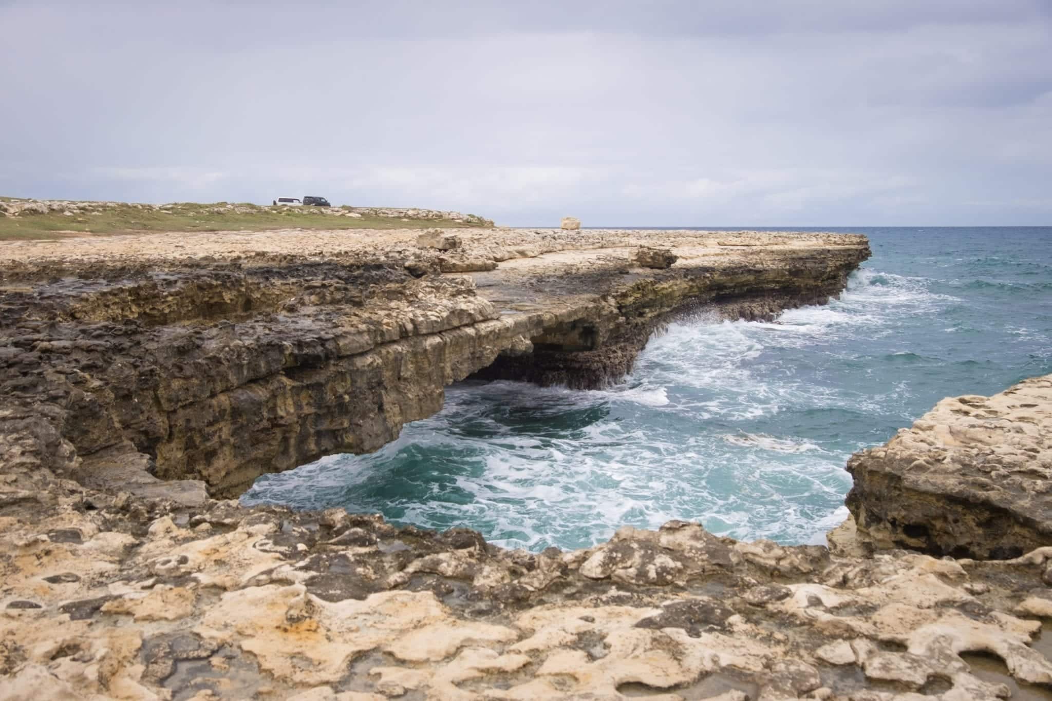 The Devil's Bridge, a rocky formation jutting out into the violent sea in Antigua. Waves are coming high, the sea and sky are both slate gray.