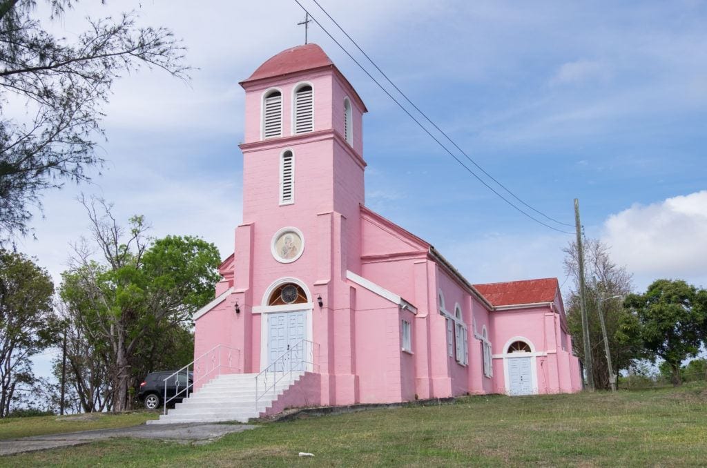 A small pink church is perched on a hill in Antigua with a soft blue and white sky in the background.