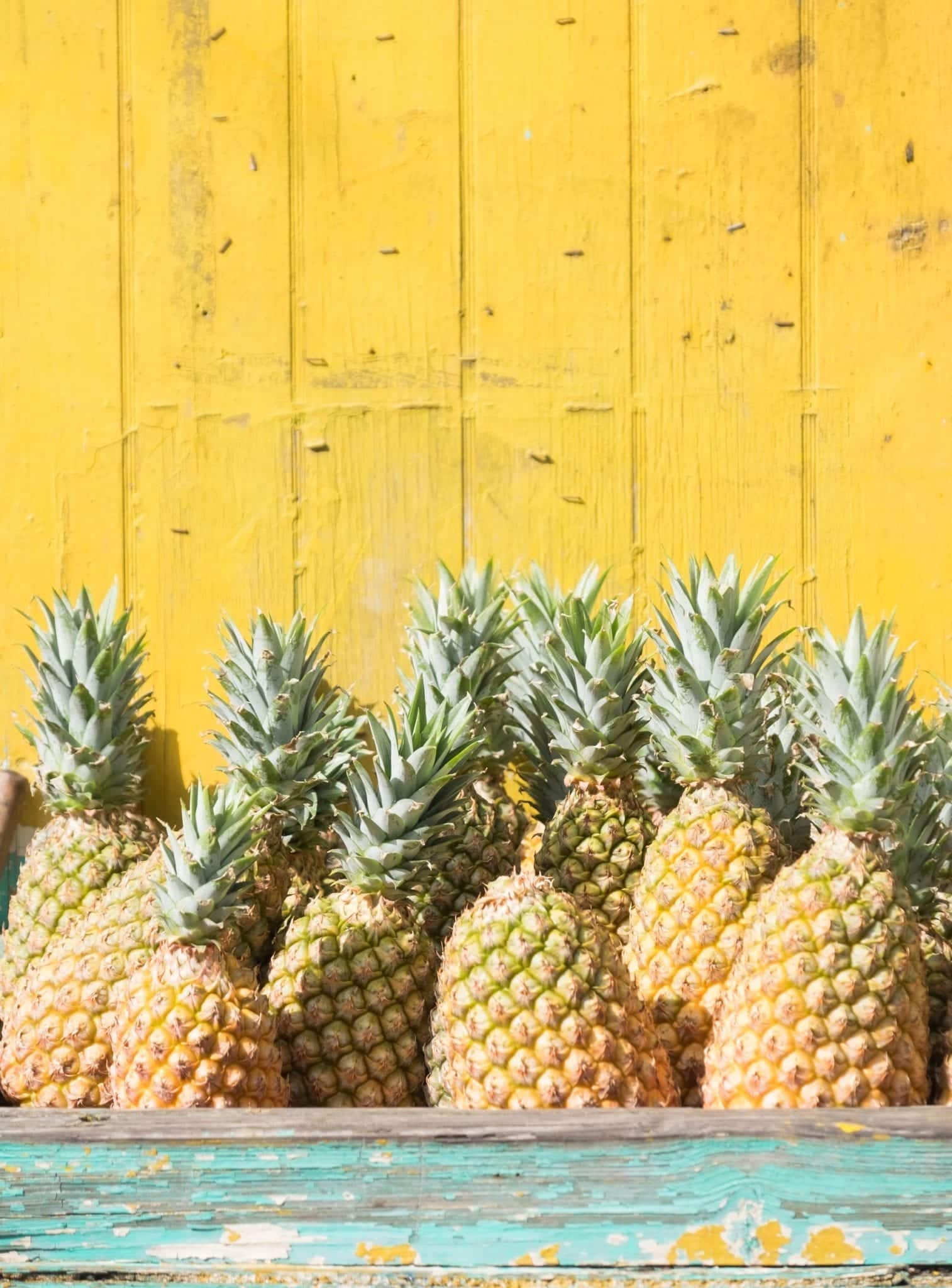 Several pineapples piled up on top of each other in a weathered turquoise wooden container in front of a yellow clapboard wall.