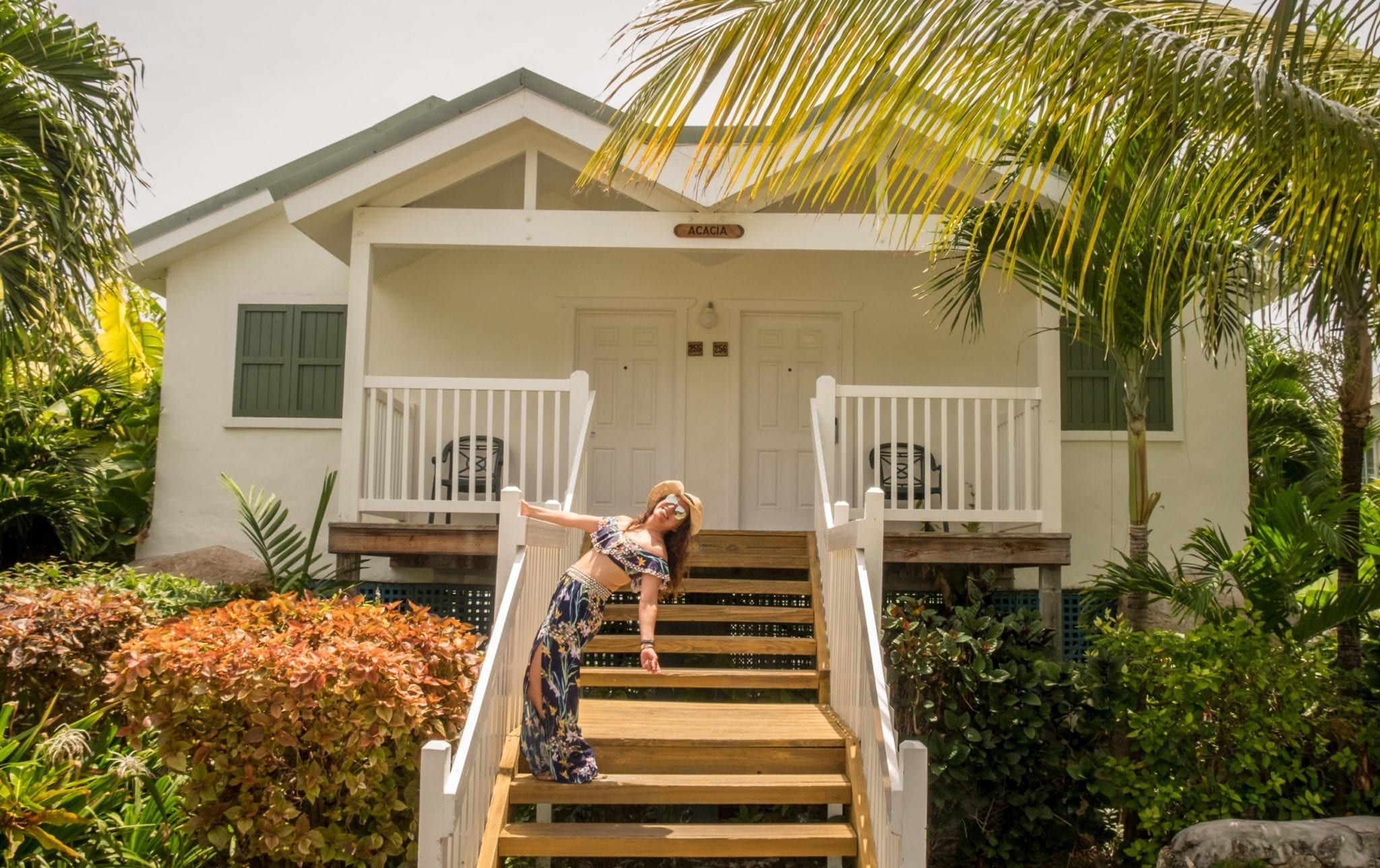 Kate posing in front of a cottage on the stairs leading up. The cottage has two doors in front and several palm trees around. Kate is holding onto the railing and arching her back so she's falling backward. She wears a straw hat, aviators, and a tropical-patterned bathing suit top that matches her genie-style pants.