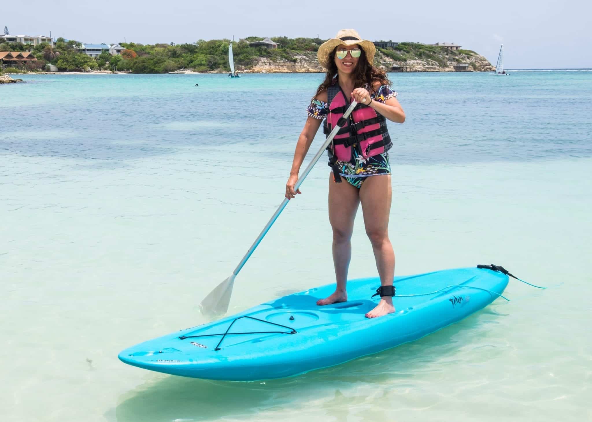 Kate paddles herself on a stand-up paddle board over turquoise water in Antigua. The board is bright blue and she wears a life vest over a bathing suit with a straw hat and aviator sunglasses.