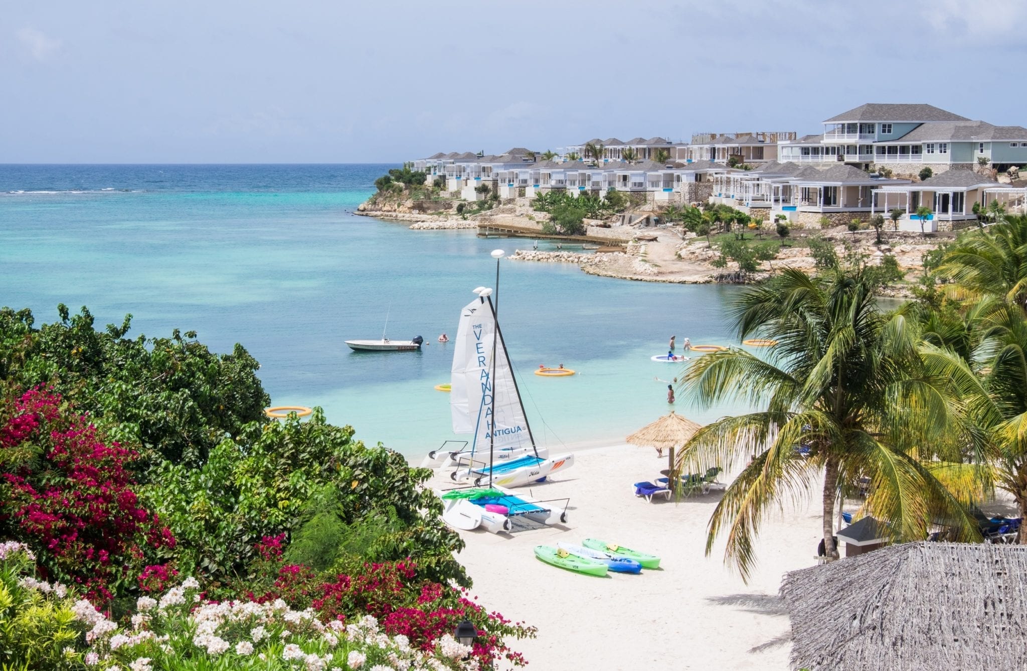 Looking down toward the beach at the Verandah Resort Antigua. You se e a turquoise ocean, white sand, a windsurfing board on the beach, palm trees on one side, and green and red plants on the other.