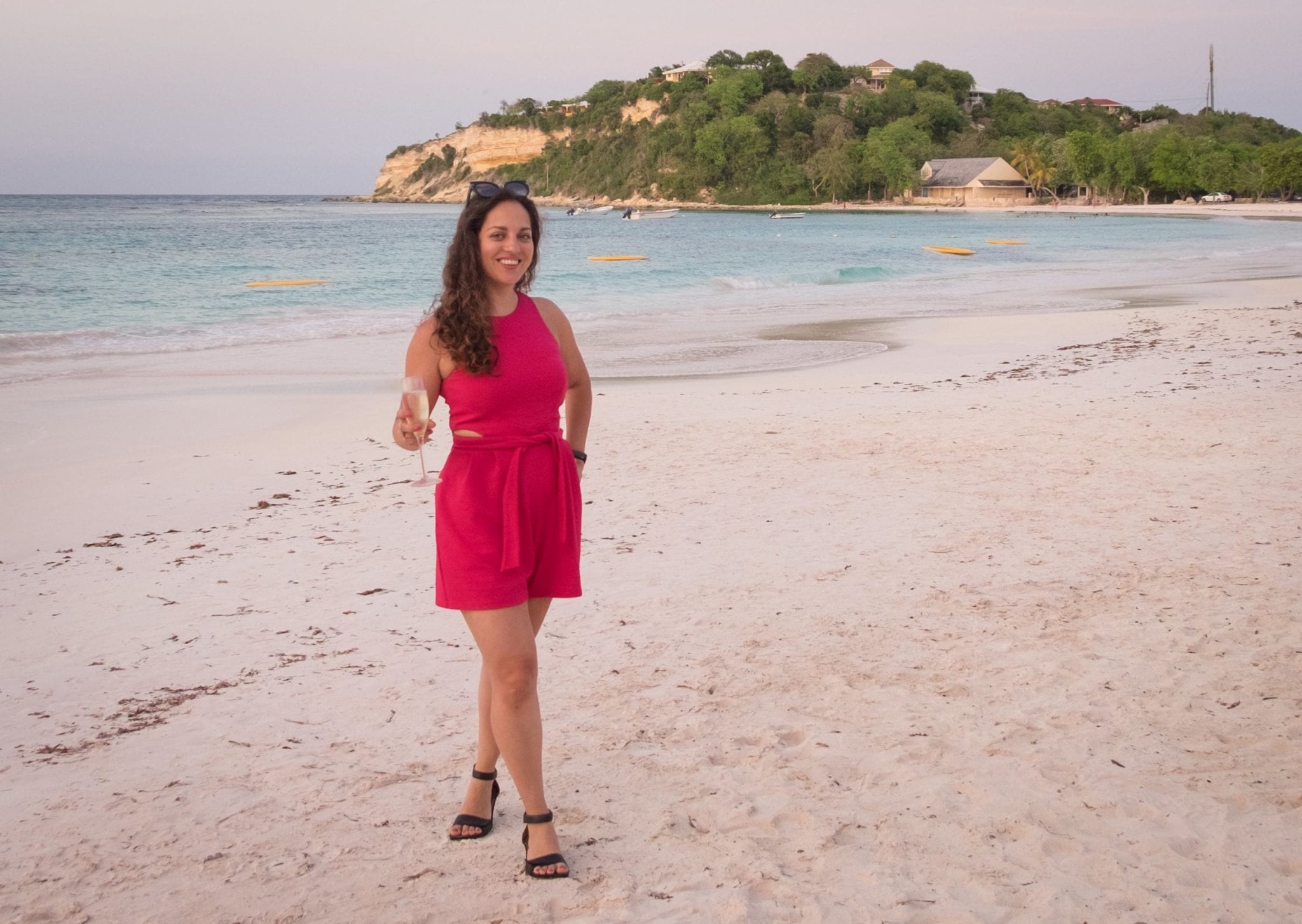 Kate wearing a hot pink romper and holding a glass of champagne, standing on a pinkish-white beach at sunset.