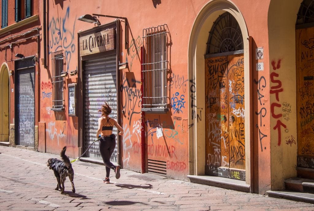 A woman in workout gear runs with her black medium-sized dog on a leash. They run past a pinkish-red wall covered with graffiti in Bologna, Italy.