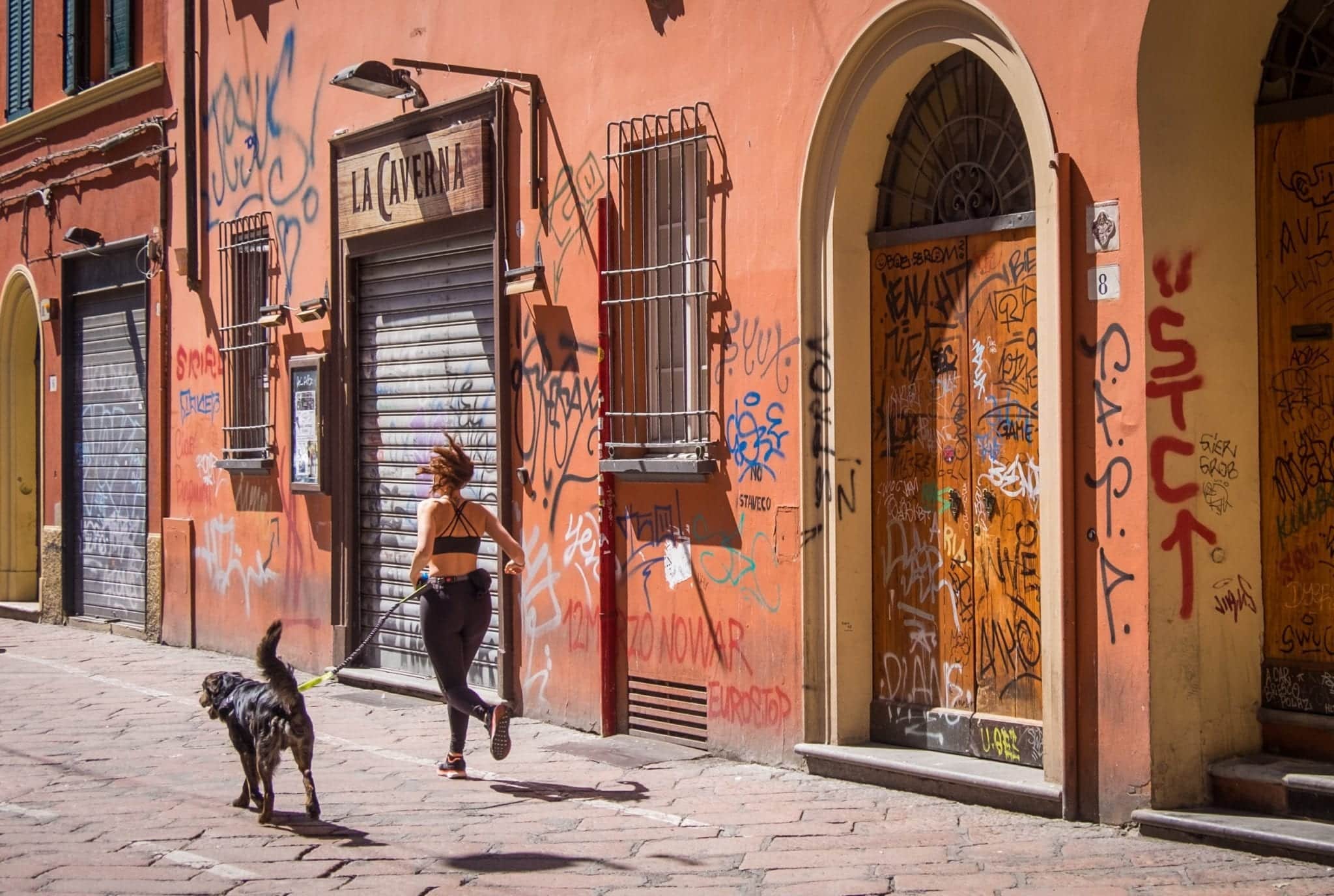 A woman in workout gear runs with her black medium-sized dog on a leash. They run past a pinkish-red wall covered with graffiti in Bologna, Italy.