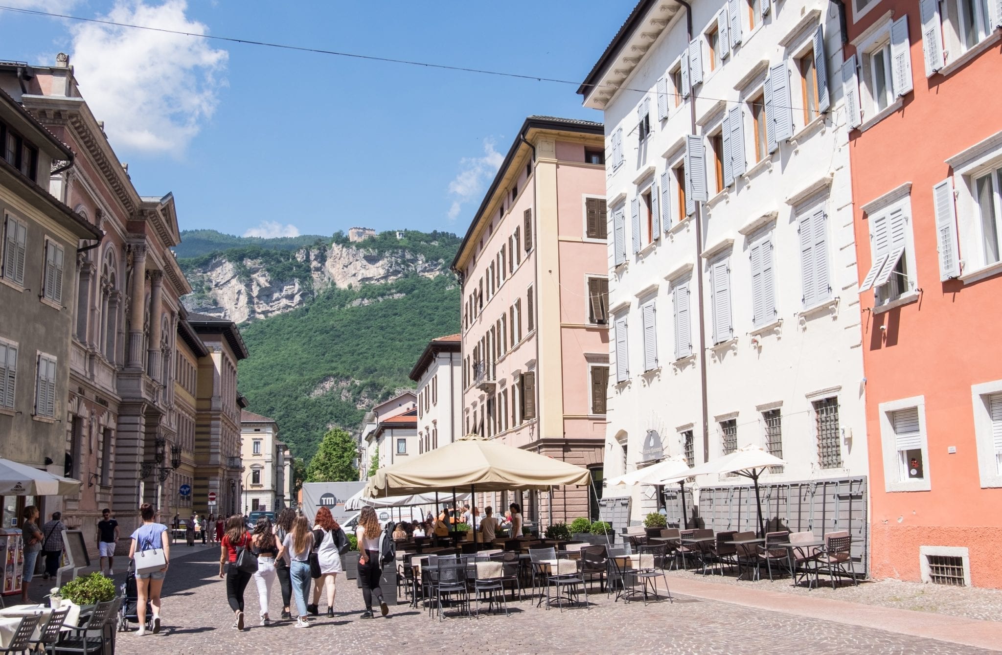 A piazza in Trento, Italy, gives way to green mountains in the background. The buildings are cream, white, and pale orange and a group of women walks together in the foreground.