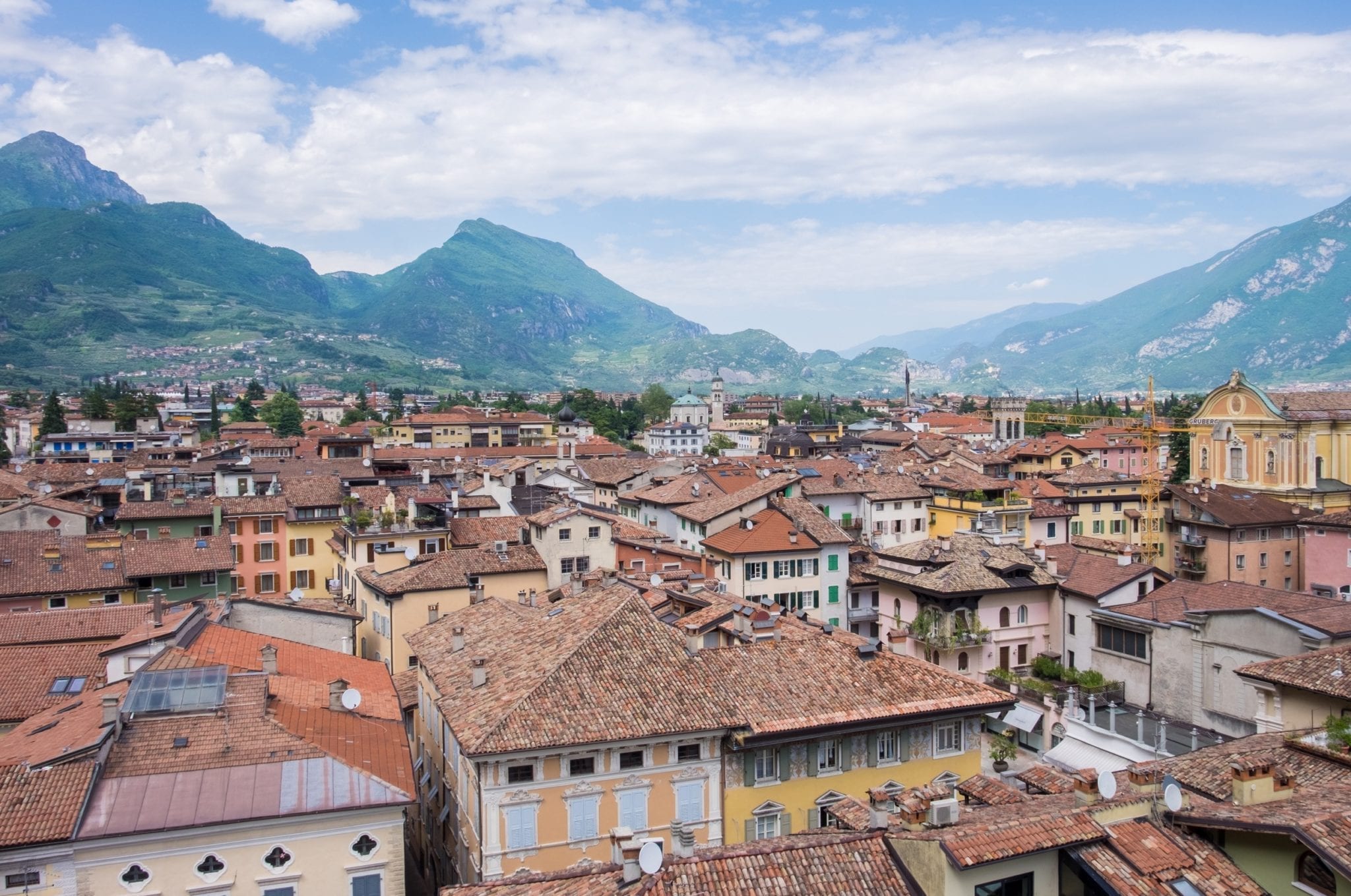 Viewing from a point above, a city of brown terra-cotta rooftops spreads over the expanse. In the distance, green mountains rise up against a blue and white-streaked sky. Riva del Garda, Italy