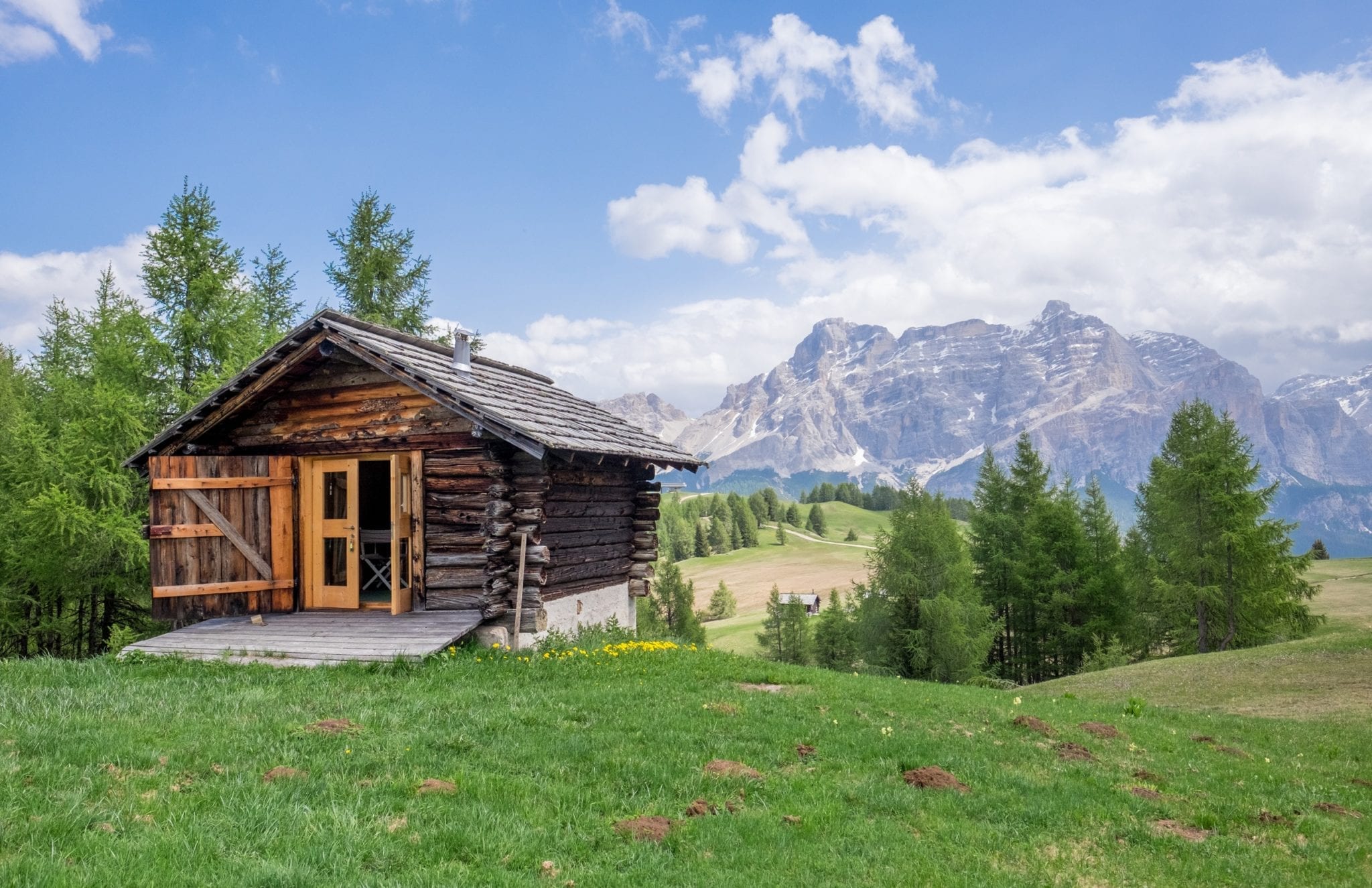 Scene from Alto Adige (South Tyrol), Italy: A wooden mountain hut, looking like a cabin, is on the left side of the screen, perched on grass; in the background, blue mountains rise up beneath a blue sky streaked with white clouds.