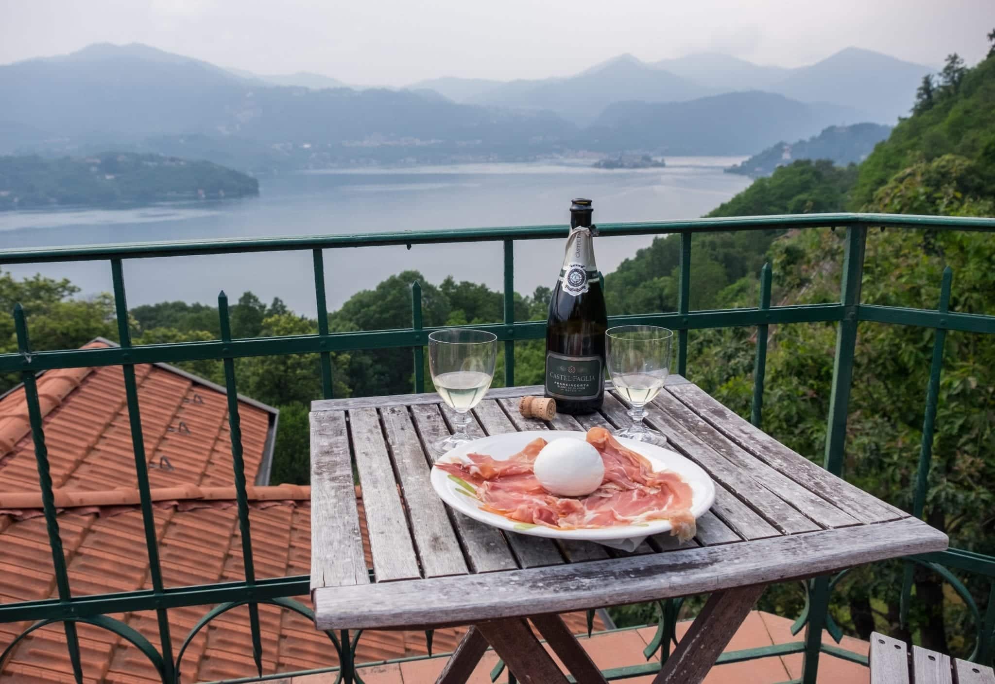 A terrace overlooks a blue and gray misty Lake Orta in the distance, mountains rising up over the lake. In the foreground there is a weathered wooden table. On it is a plate covered with prosciutto and a ball of burrata cheese; behind it are a bottle of Franciacorta sparkling white wine and two goblets filled with the wine.