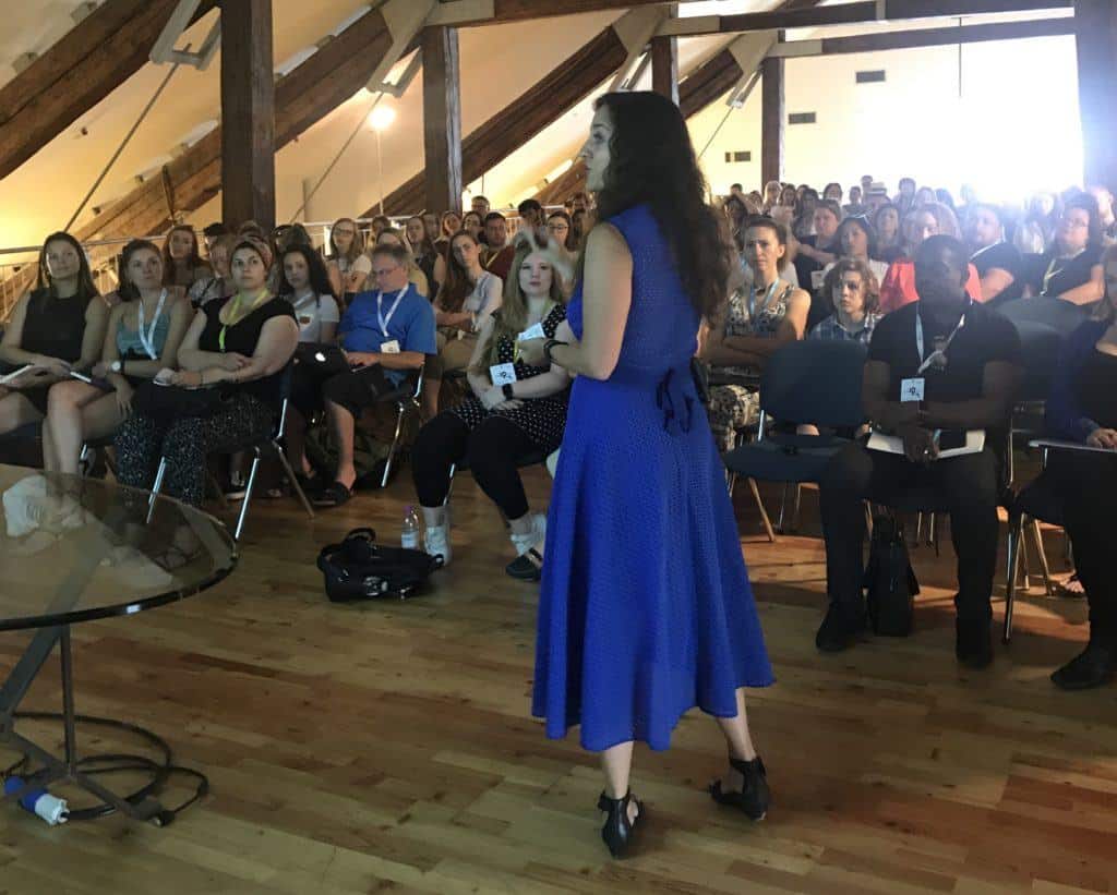 Kate is wearing a long bright blue dress and speaking toward a packed room with more than 60 people watch her giving a presentation.