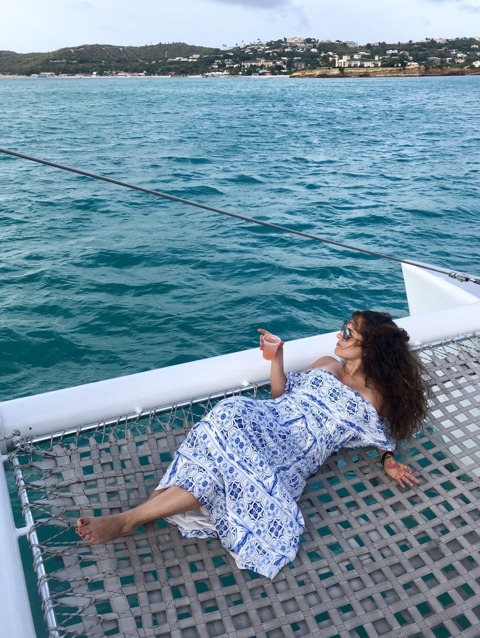 Kate lying down on a net on the end of a catamaran, wearing an off the shoulder white and blue patterned dress, hair long and curly, leaning on her elbows, holding a plastic cup of rum punch in one hand, and staring off into the distance -- turquoise sea and green land in the distance.