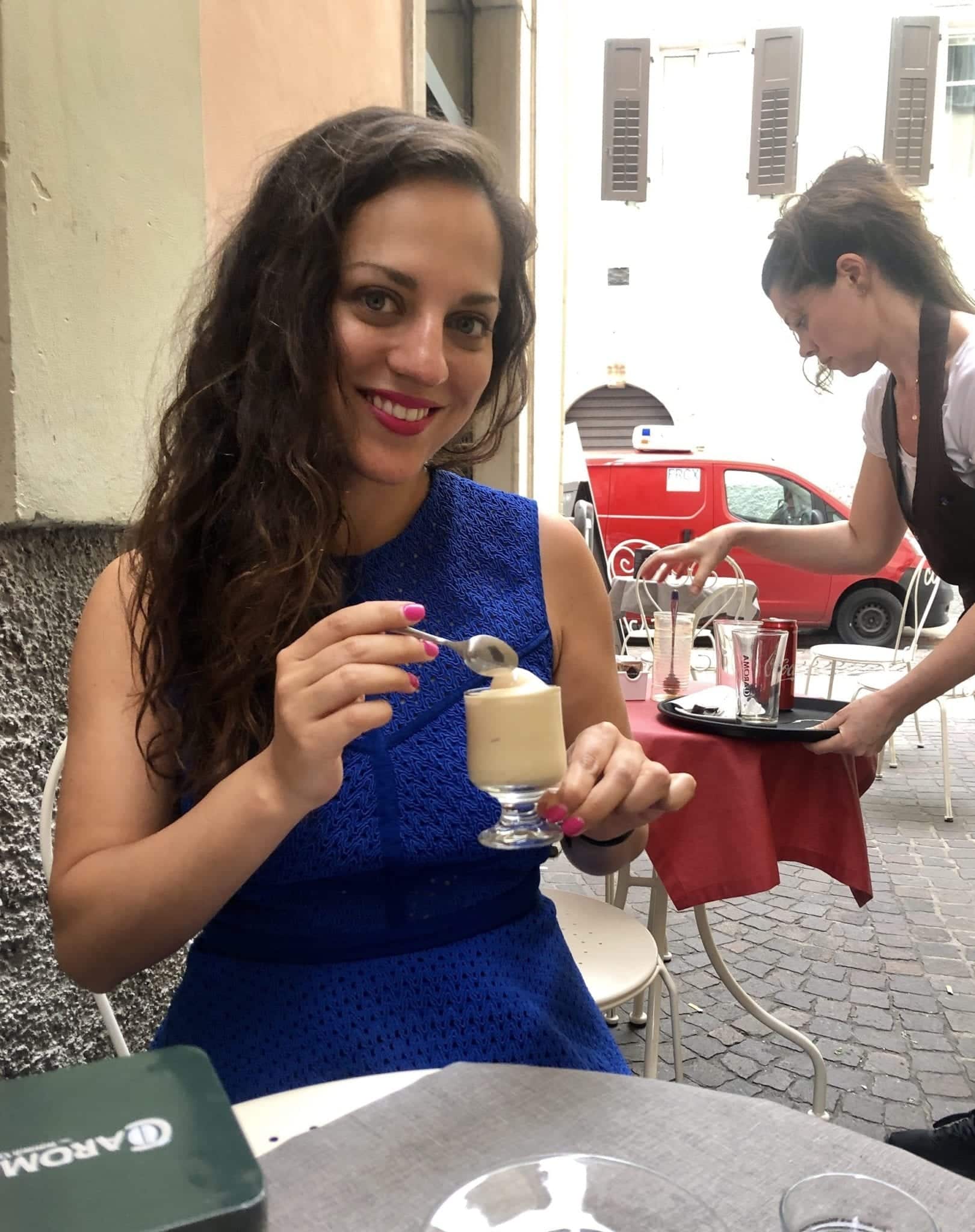 Kate wears a royal blue Pinko dress that goes up to her neck and has a transparent lacy fabric. Her hair is down a curly and she holds a cafe crema (soft serve espresso) in her hand. She is sitting at a sidewalk cafe in Trento.