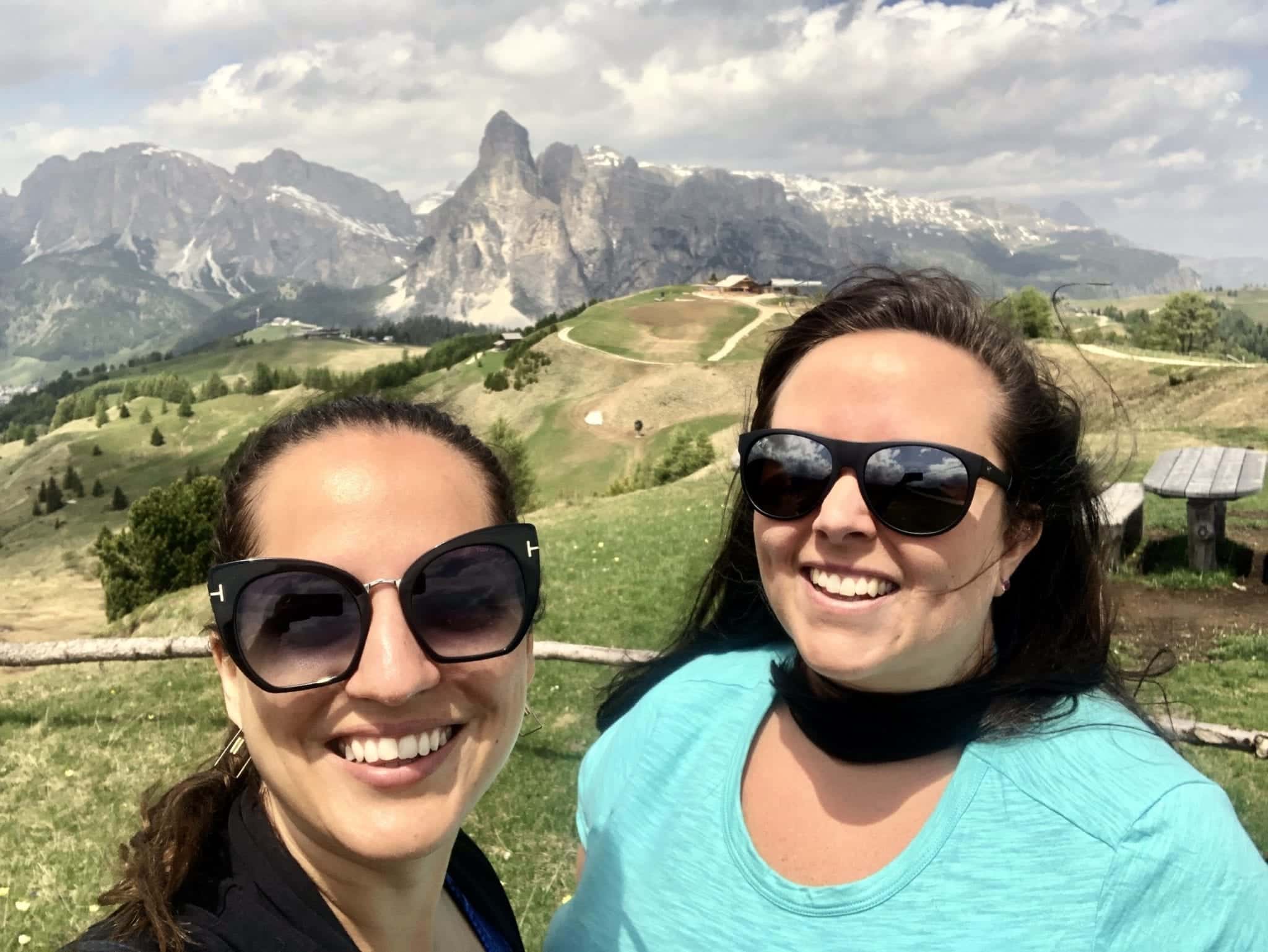 Kate and Cailin take a selfie wearing sunglasses in the Italian Dolomites. Behind them is a row of jagged mountains in the distance. There is a dramatic cloudy sky.