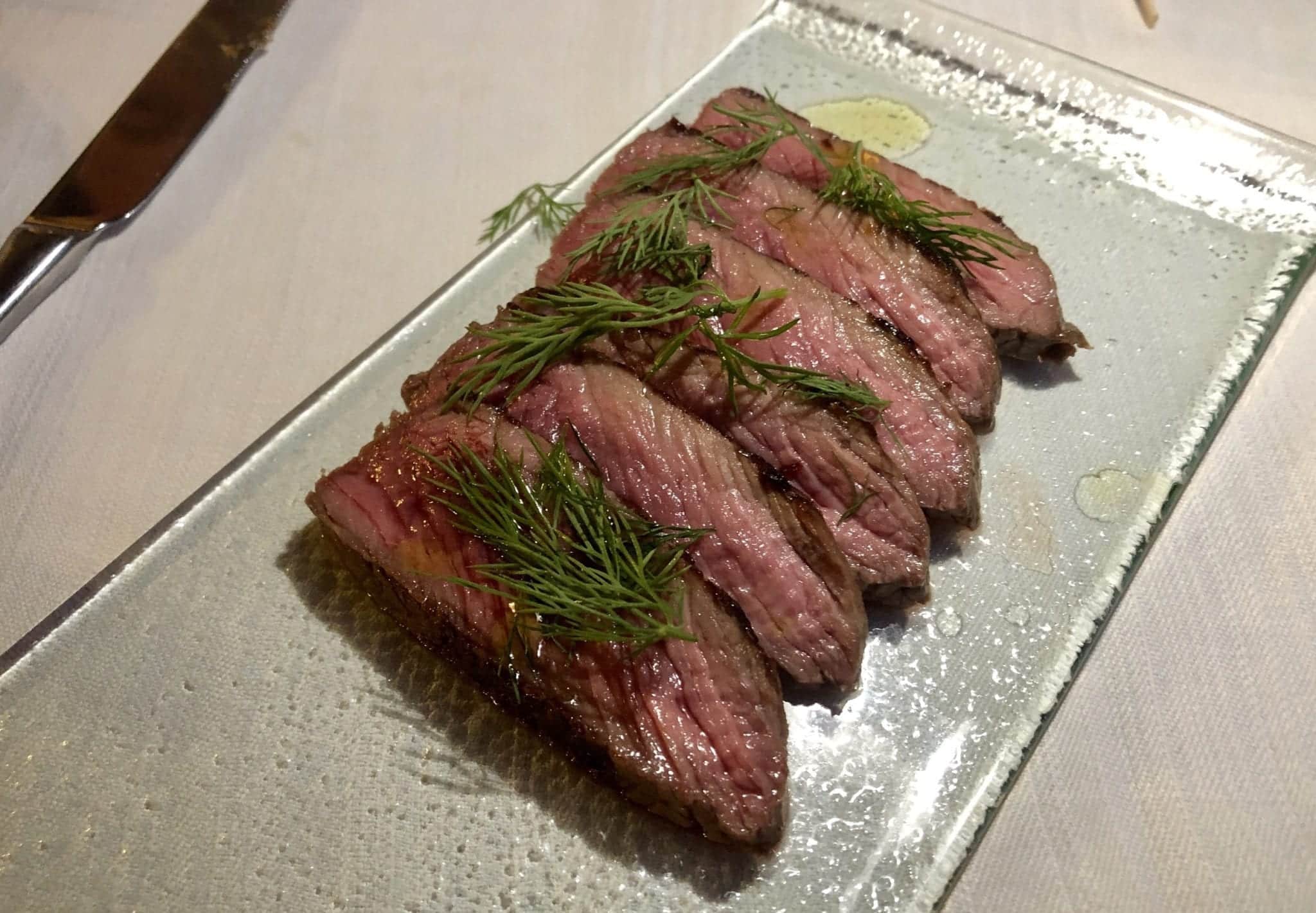 Tagliata di Manzo -- thin slices of filet mignon, topped with dill and served medium rare, sitting on a clear glass plate and a white tablecloth.