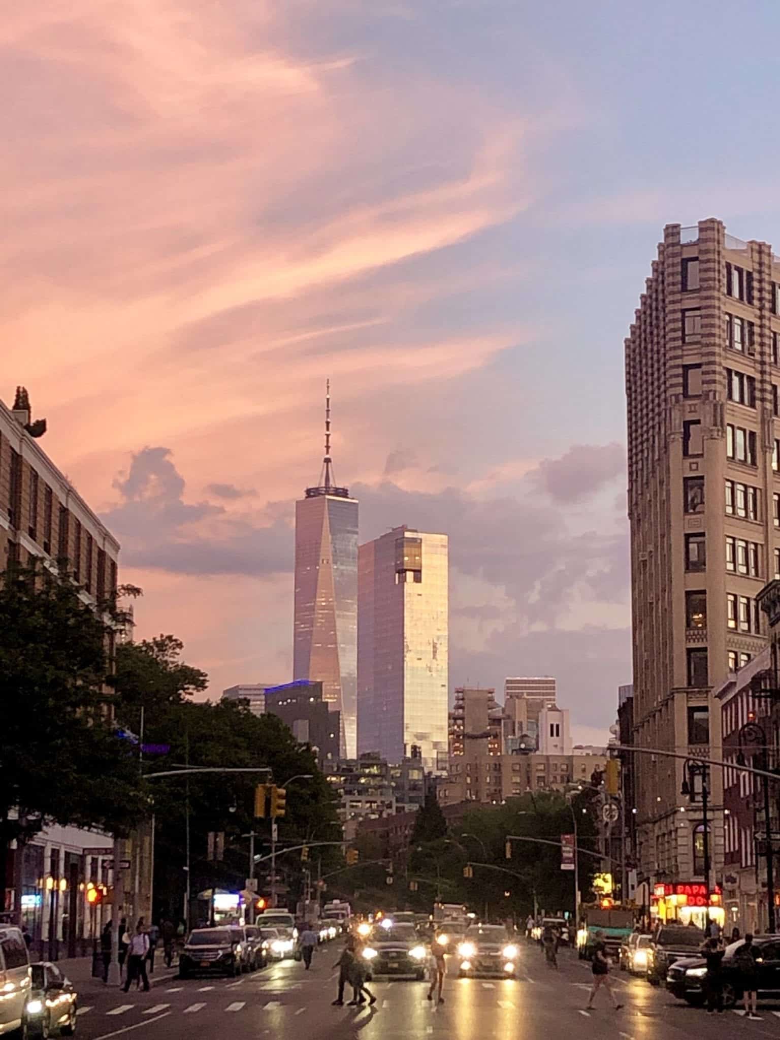 The One World Trade Center building in New York is illuminated by a pink and blue sunset streaked with clouds. The view is facing straight down Sixth Avenue and there are a few sets of headlights facing the camera.