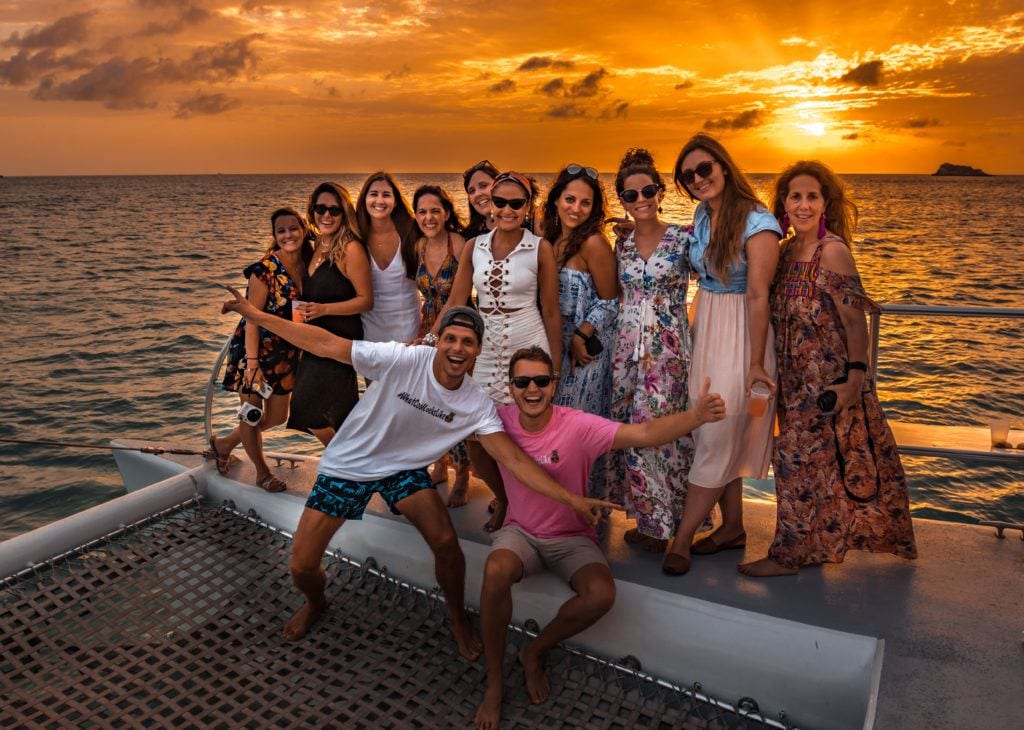 A group of friends posing on a catamaran with a bright orange sunset behind them.