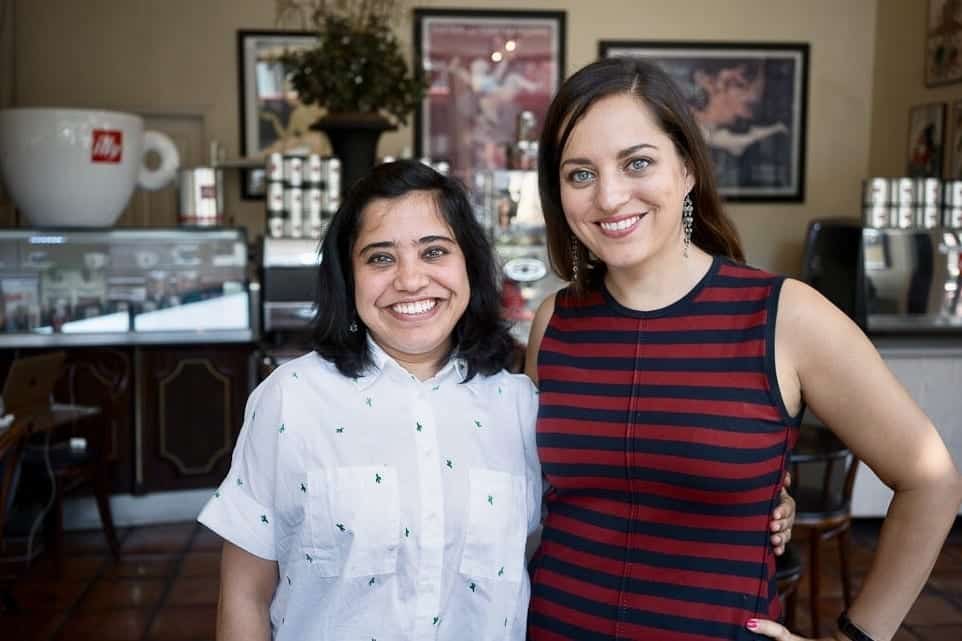 Kate in a red-and-navy-striped dress arm and arm with her friend Paroma, wearing a white button-up shirt, standing in a coffeeshop in San Francisco in front of a display case with a giant Illy coffee cup on top.