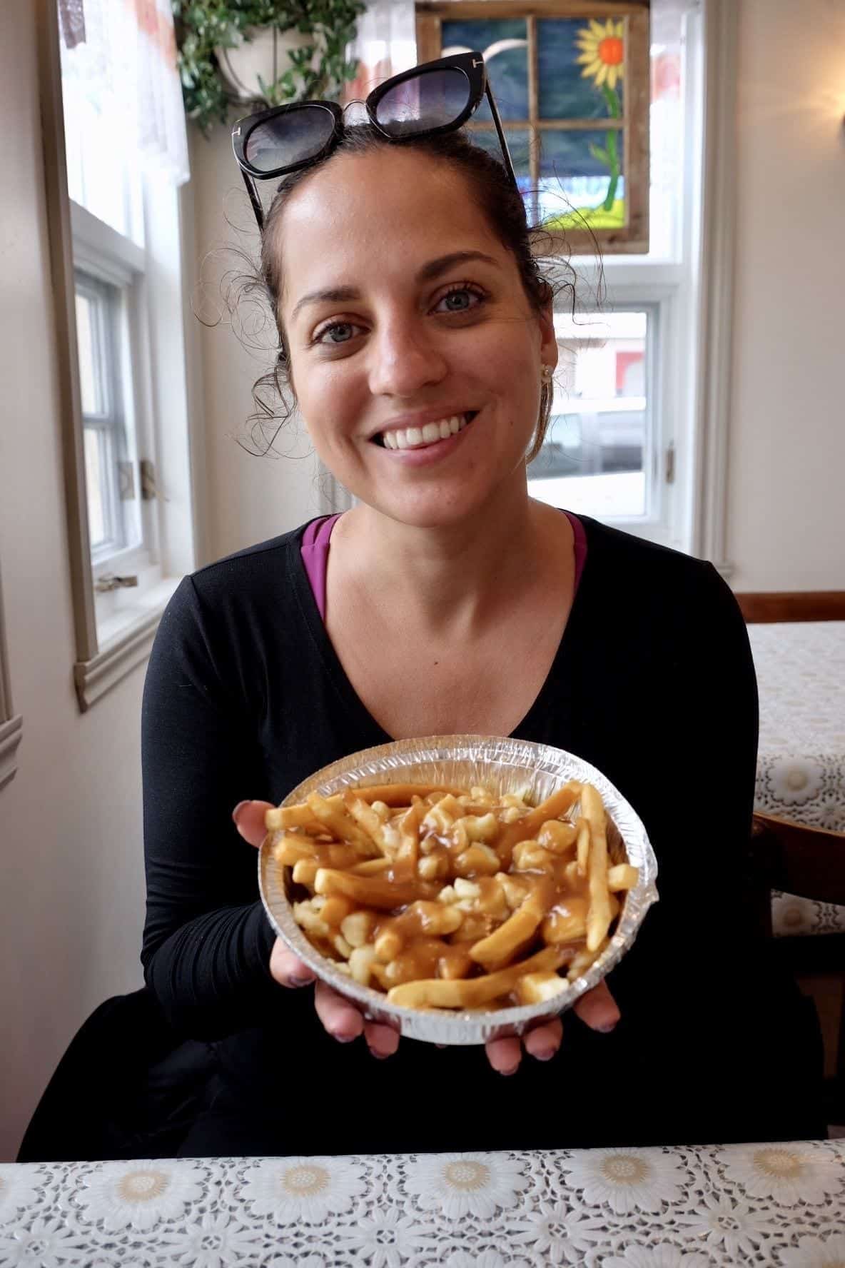Kate holds a bowl of poutine in her hand and smiles.