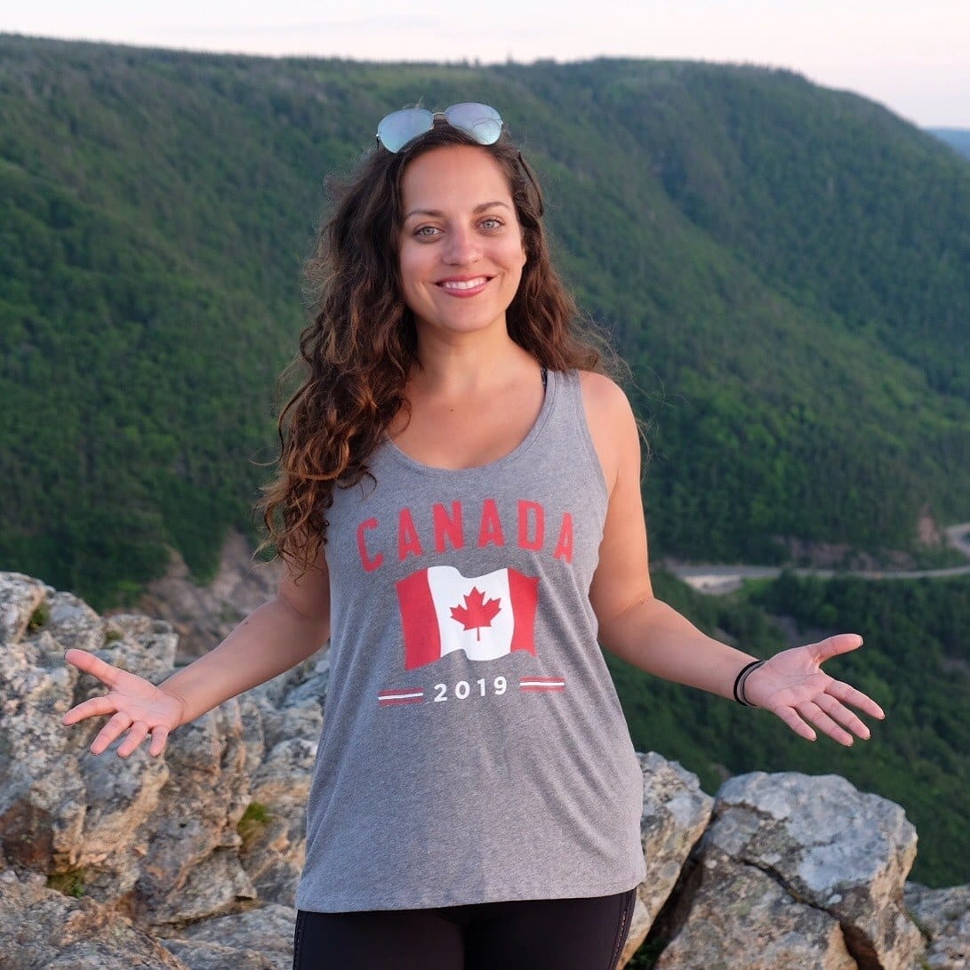 Kate wears a tank top that reads "Canada" with a Canadian flag on it and stands above Cape Breton Highlands National Park, mountains and winding roads behind her.