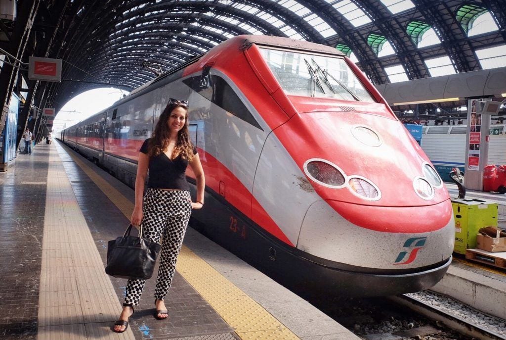 Kate stands wearing a black shirt and black and white patterned pants, holding a black purse, in front of a modern Frecciarossa train at Milan train station. The train is sleek and is silver and red with a long angled nose in front.