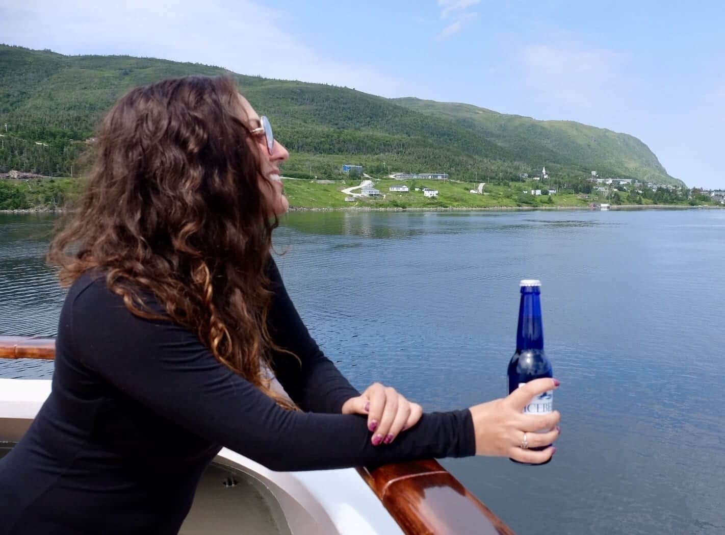 Kate leans over the edge of a ship, holding a cobalt blue Iceberg Beer bottle in her hand and looking over the glassy bright blue Bonne Bay in Newfoundland.