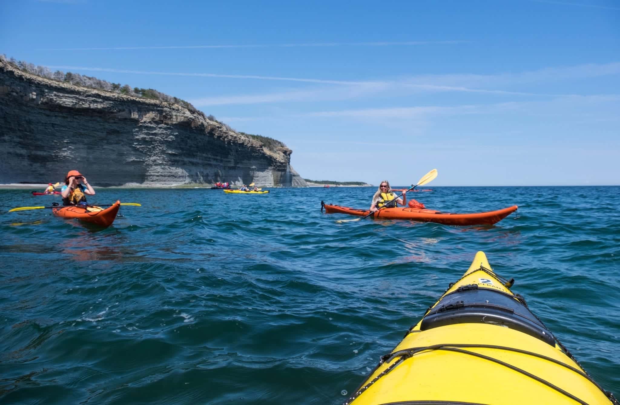 Shot from the front of a yellow kayak, you see several kayakers in bright blue water and cliffs in the background.