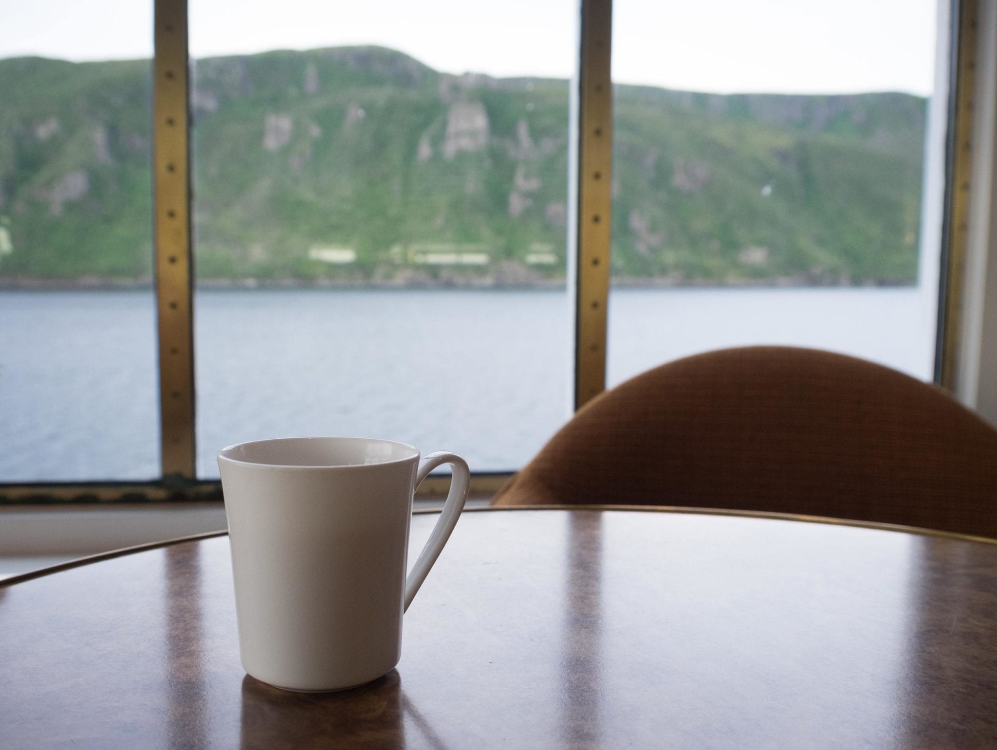 A cup of tea sits on a table in front of windows revealing the fjord at Bonne Bay, Newfoundland.