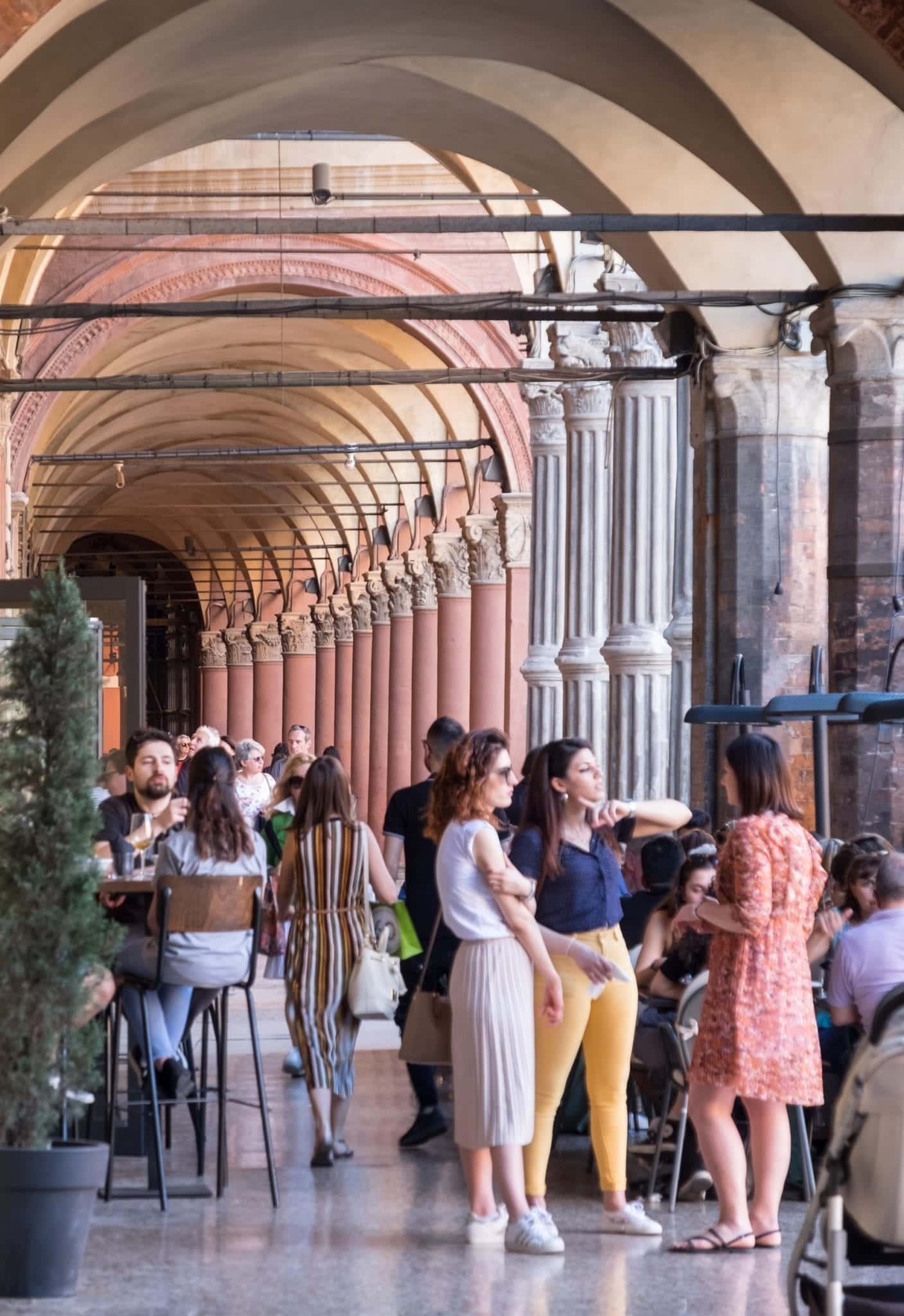 Three women stand talking beneath porticoes stretching through the background.