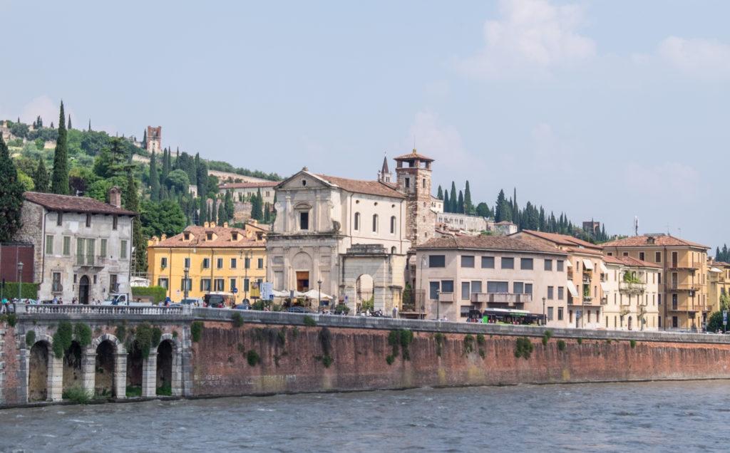 The riverbank in Verona, Italy, with cream-colored buildings and tall cypress trees.