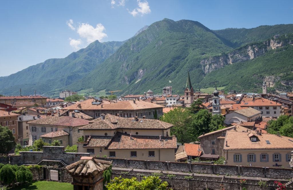 A view of the terra cotta roofs in the old town of Trento, Italy, with green mountains in the background underneath a blue sky with white spotted clouds.
