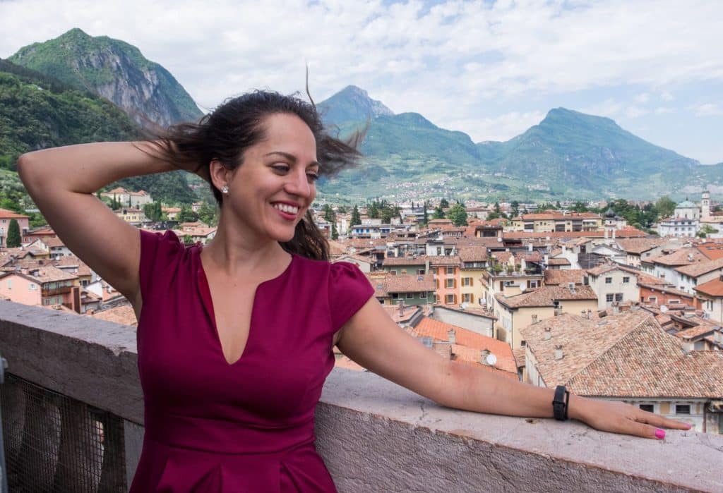 Kate smiles with her hand behind her head and wears a red dress and stands on top of a tower in Riva del Garda, Italy, overlooking terra cotta roofs, pastel buildings, with jagged mountains and a white and blue streaked sky in the background.