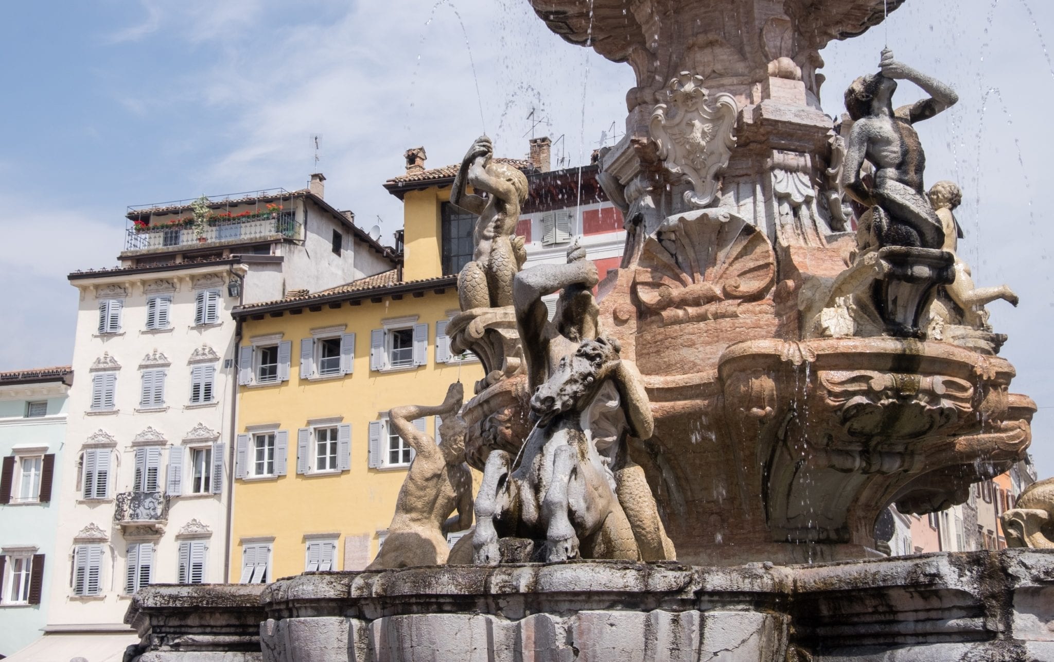 A fountain covered with nymphs spurts out water next to yellow and white buildings in Trento, Italy.