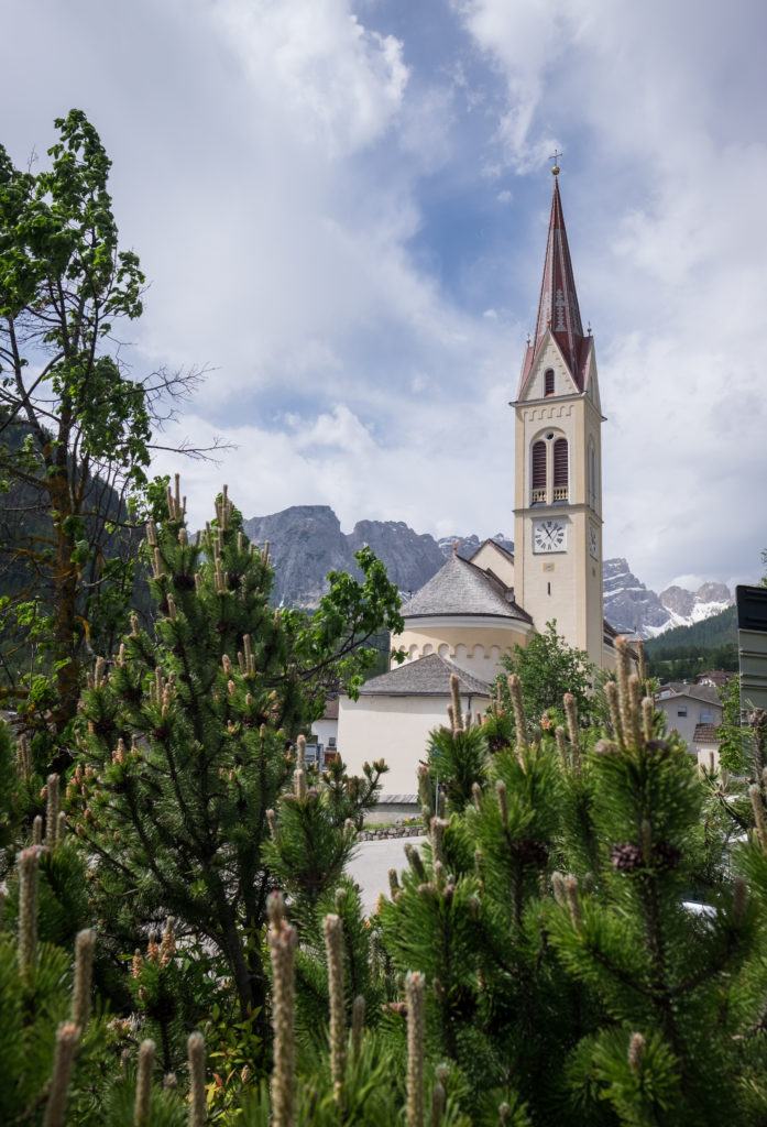 A church steeple pokes out of an Italian mountain landscape in the Dolomites. The church is surrounded by forest.