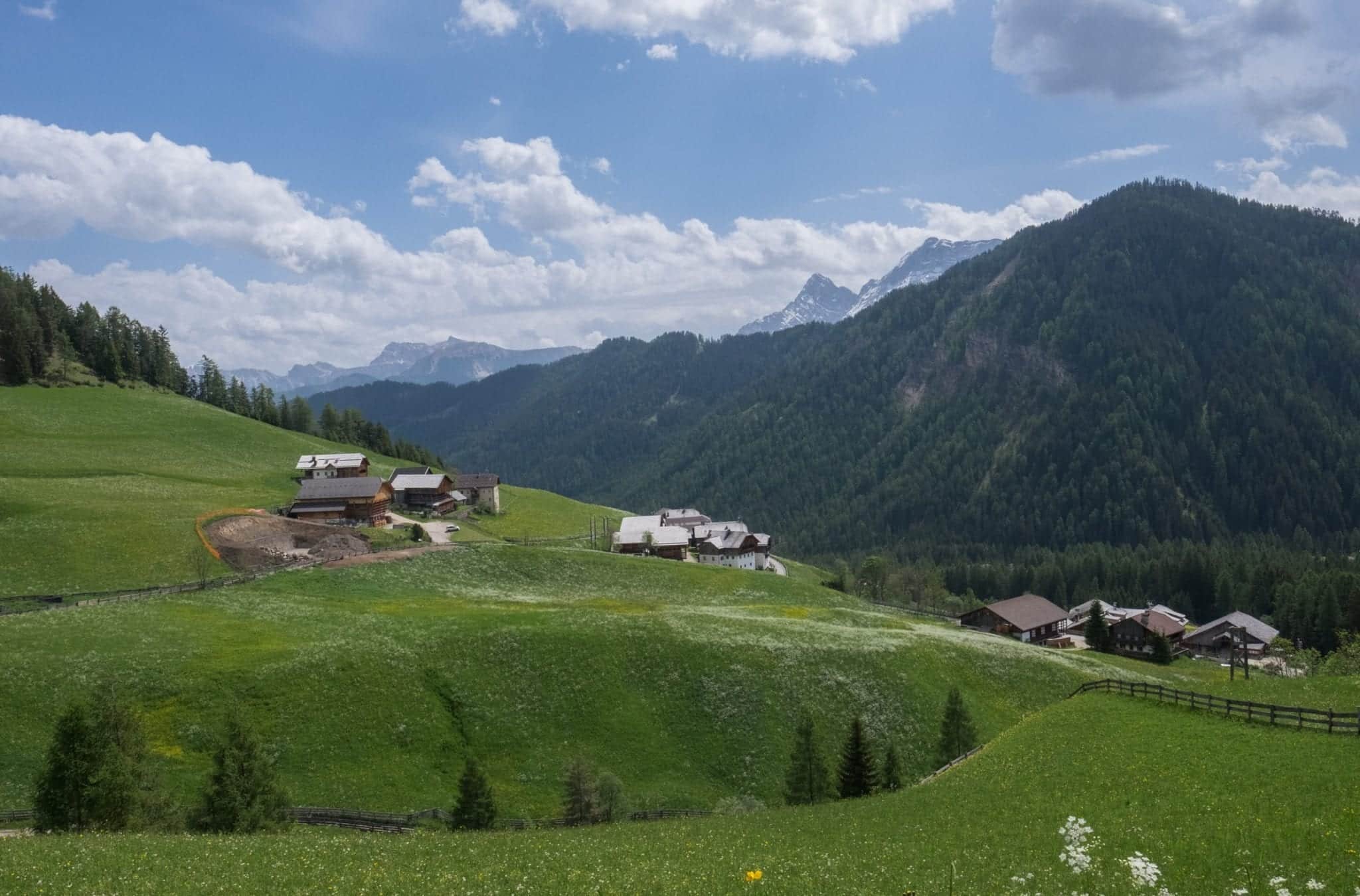 A view of the Dolomites with mountains in the distance and tiny Alpine huts on green hills in the foreground.