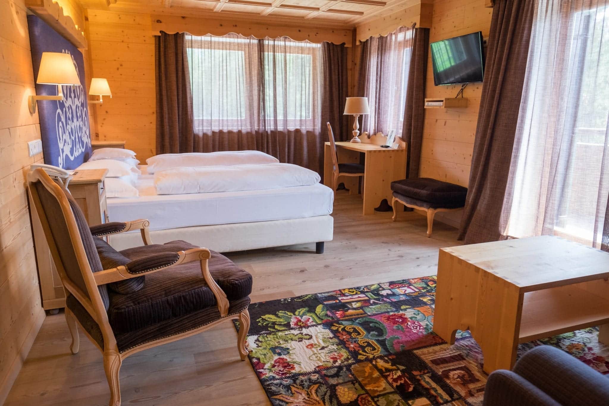 A hotel room with a white double bed, modern wooden furnishings, and a multi-colored traditional rug on the ground.