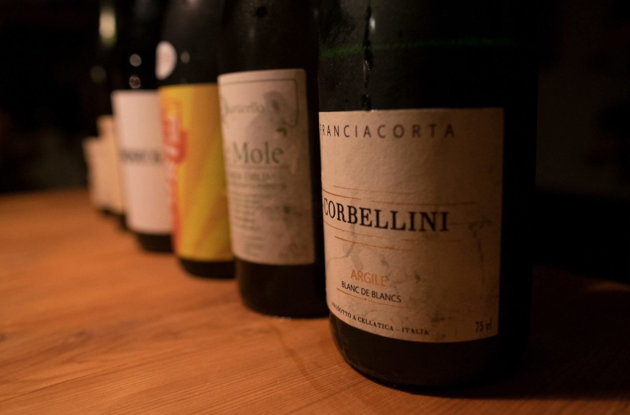 Several wine bottles lined up in a row in dim light, leading with a Franciacorta.