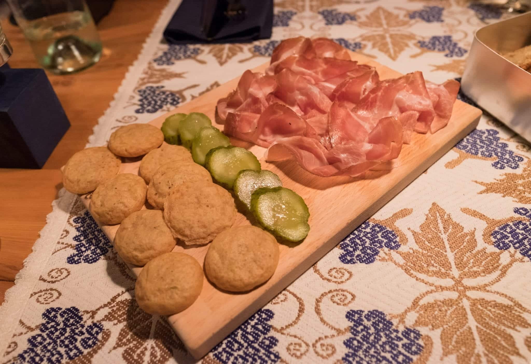 A platter covered with round pieces of fried bread, pickles, and pink feathery speck.