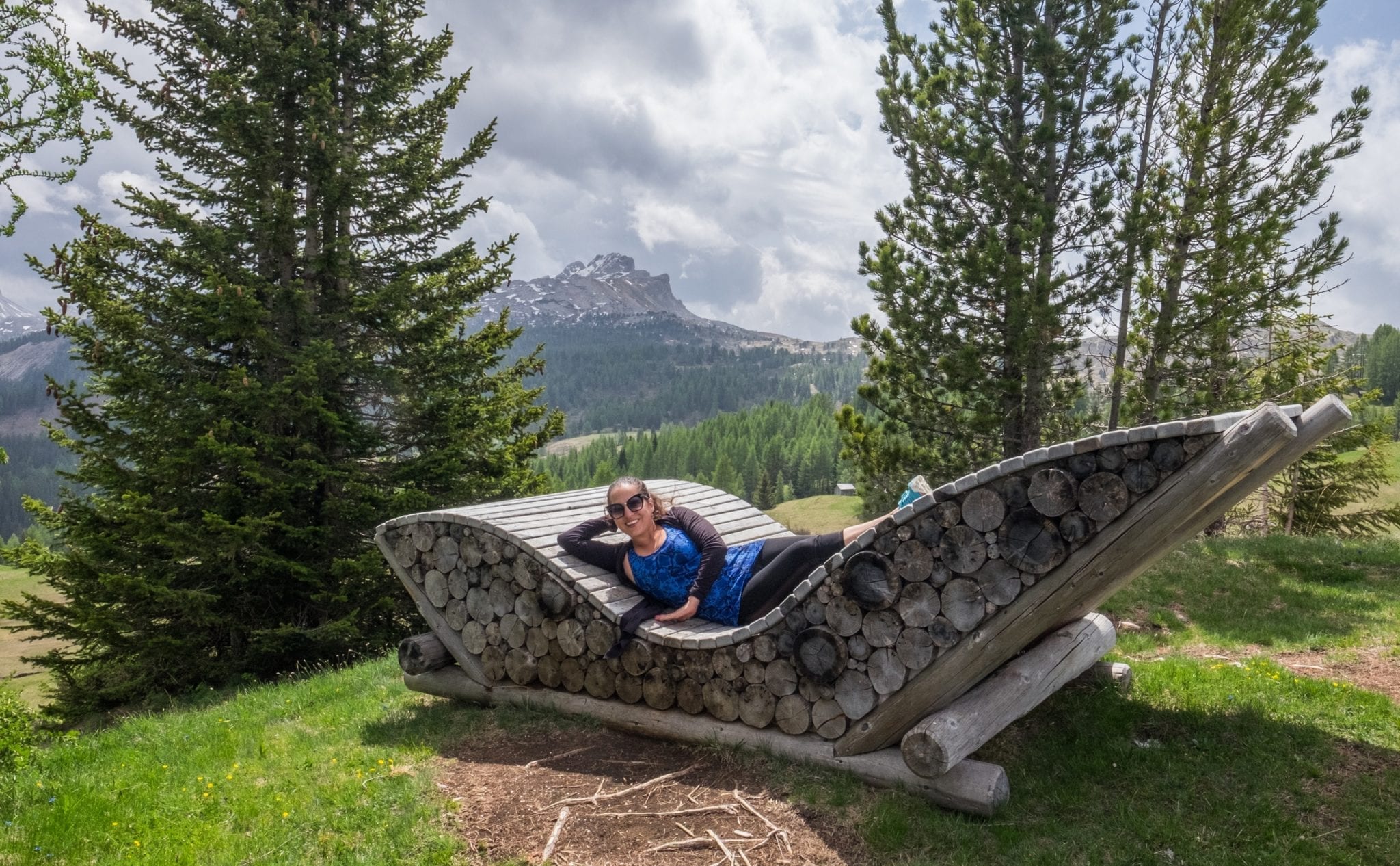 Kate stretches out on a sun lounger in the Dolomites -- it looks like a large chair, and the bottom is made of stacked wood. Trees and a Mountain View are in the distance.