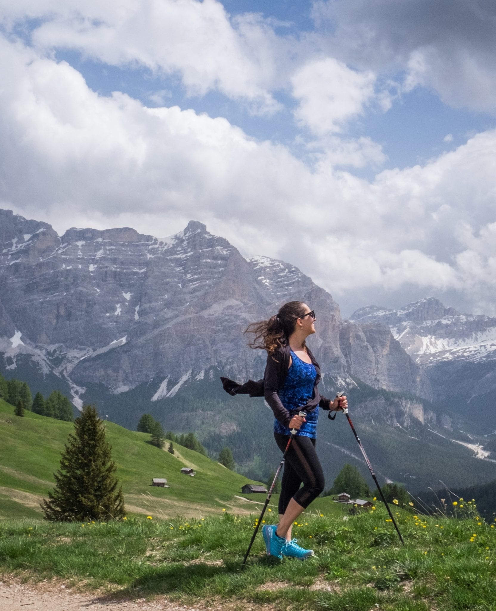 Kate stands on a hill in the Dolomites, holding hiking poles in her hands, illuminated by purple-gray mountains behind her and a cloudy blue and white sky.