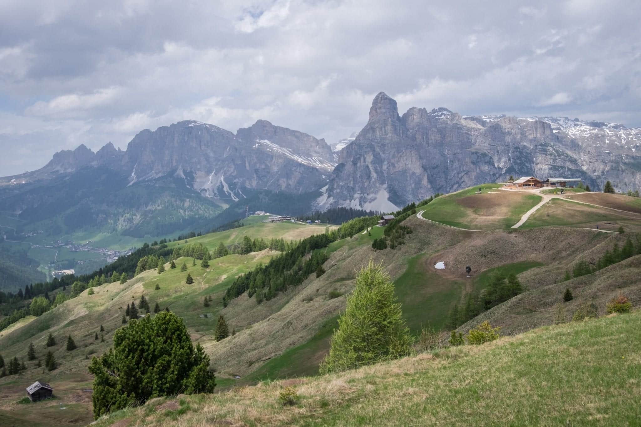 View of the Dolomites: jagged gray peaks in the background and mossy green hills in the foreground. A gray cloudy sky overhead.